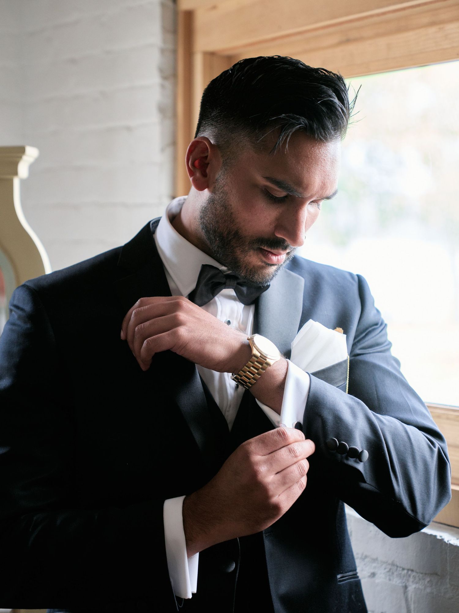 A groom in a blue tuxedo adjusts his cufflinks while getting ready near a window during wedding preparations.