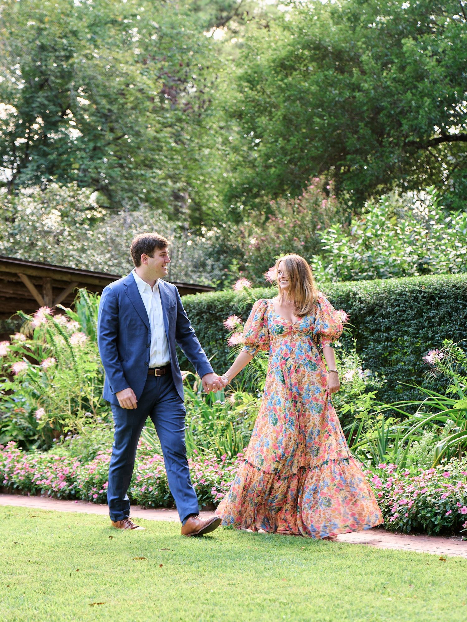 Couple playfully holds hands in garden setting with woman wearing floral maxi dress.