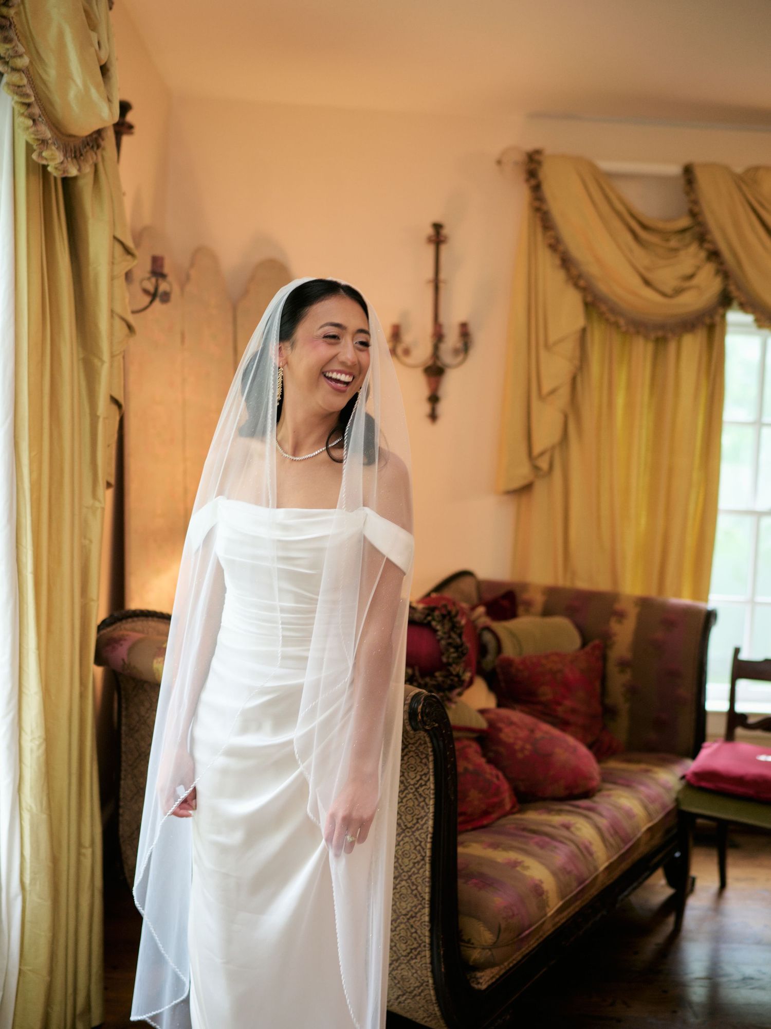 A smiling bride in a white gown and veil stands in an elegantly decorated room with gold drapes.
