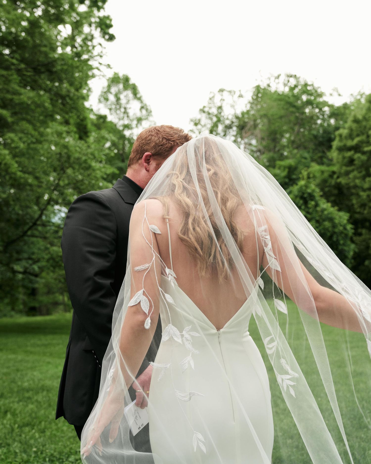 A couple in wedding attire stands together in a lush green garden setting with a flowing white veil catching the breeze.