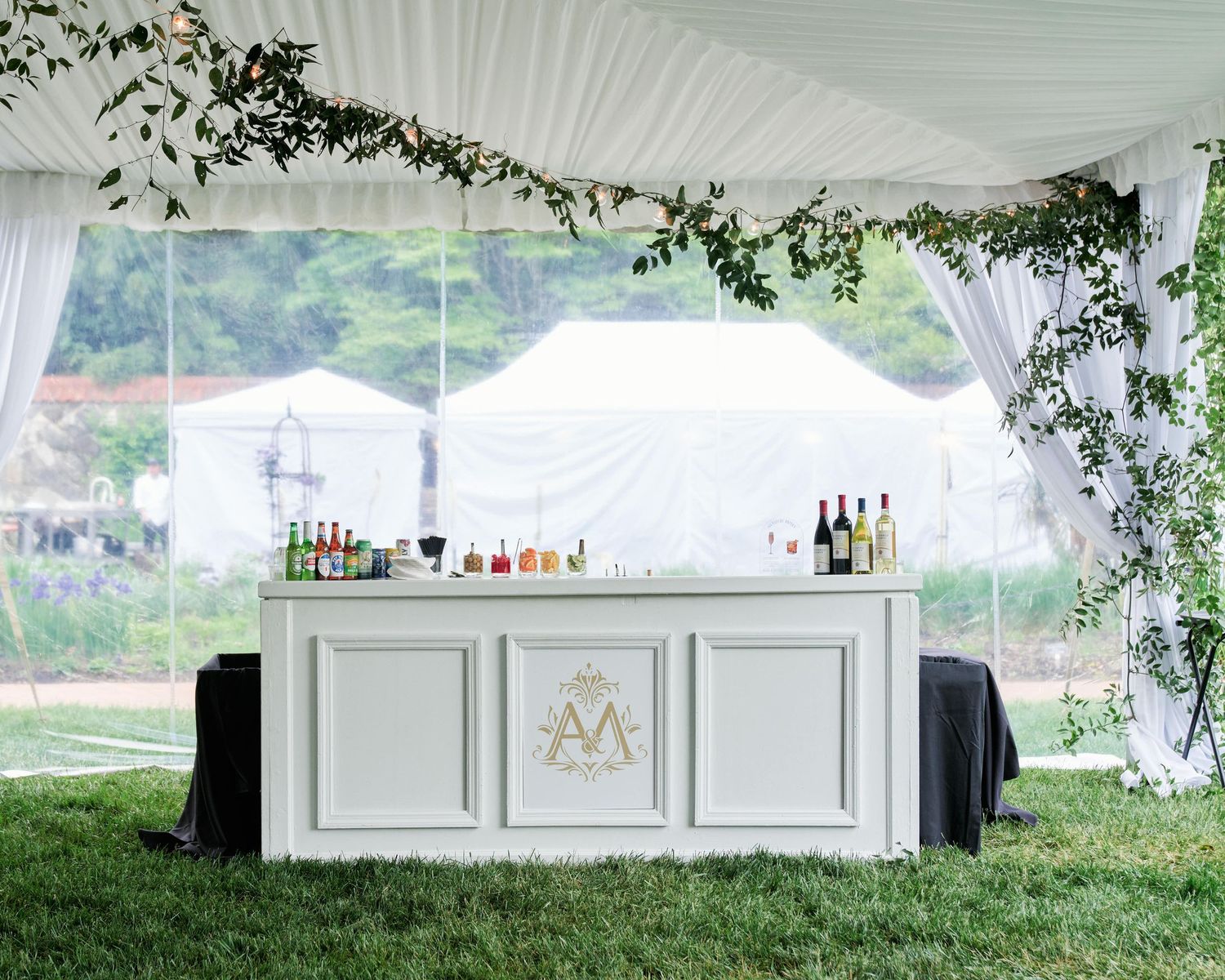 Elegant white wedding bar setup under tent with greenery garland and monogram detail on outdoor lawn.