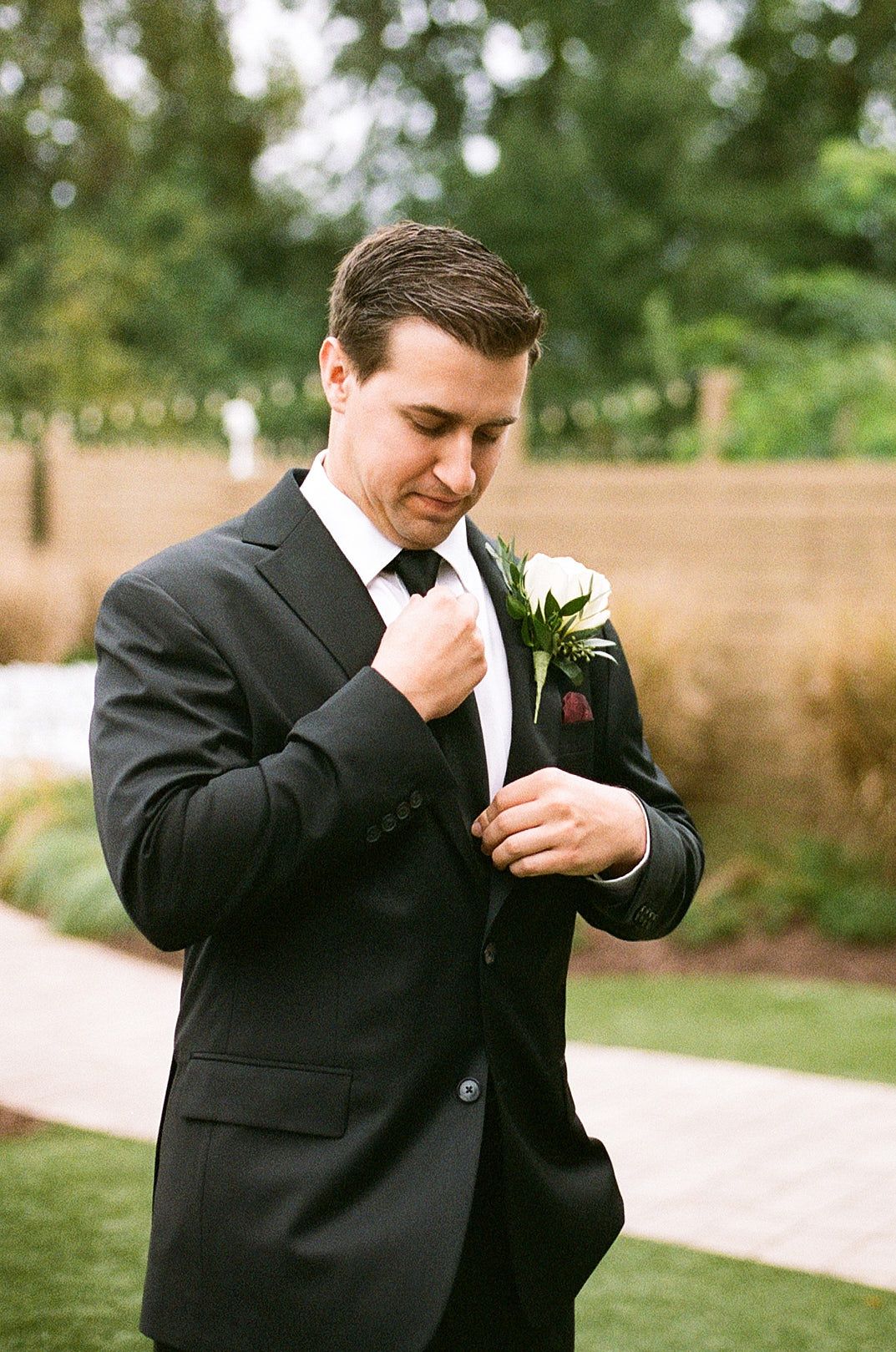 A groom in a classic black tuxedo adjusts his bowtie while wearing a white boutonniere on his lapel at an outdoor wedding.