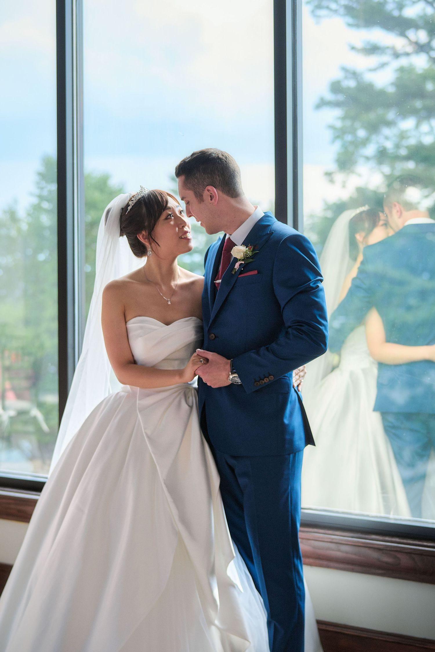 A bride in a white gown and groom in navy suit share an intimate moment by large windows overlooking greenery.