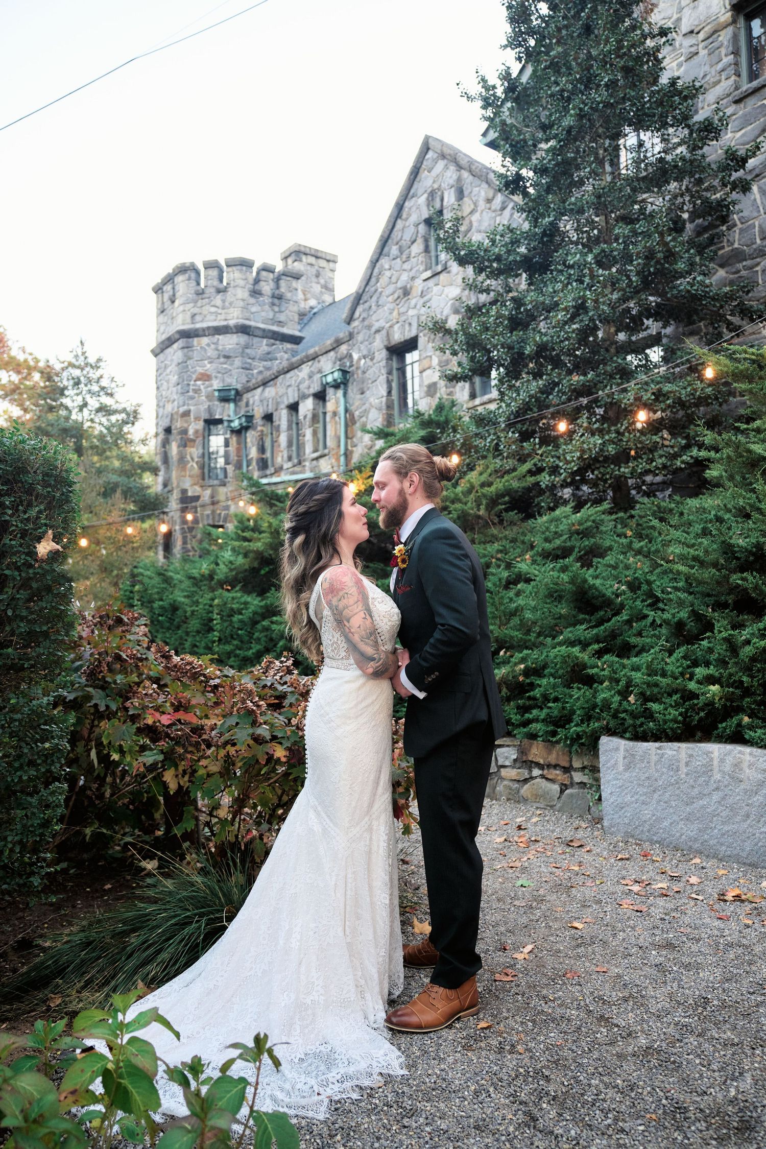 A couple shares an intimate moment outside a castle-like stone building surrounded by evergreen trees.