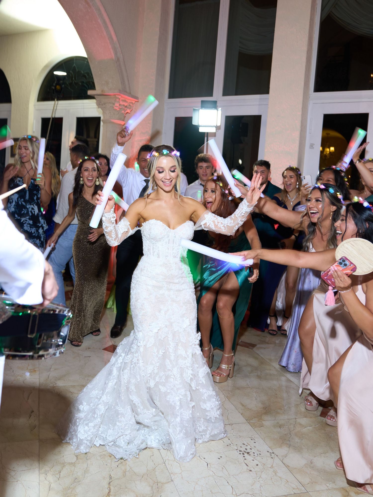 Bride in white lace gown celebrates with guests waving light sticks during wedding reception.