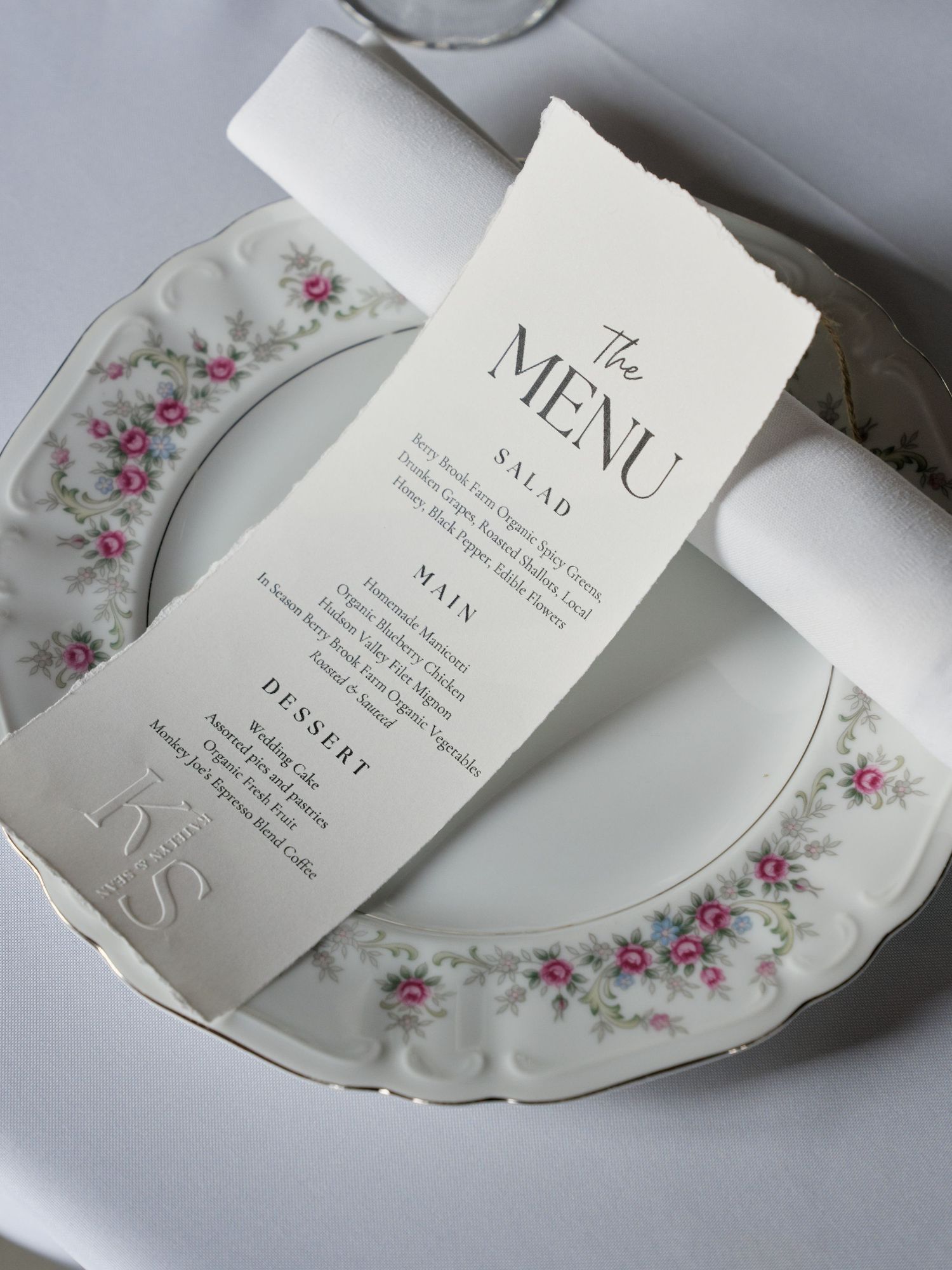 A vintage floral china plate with pink roses holds an elegant wedding menu card on a white tablecloth.