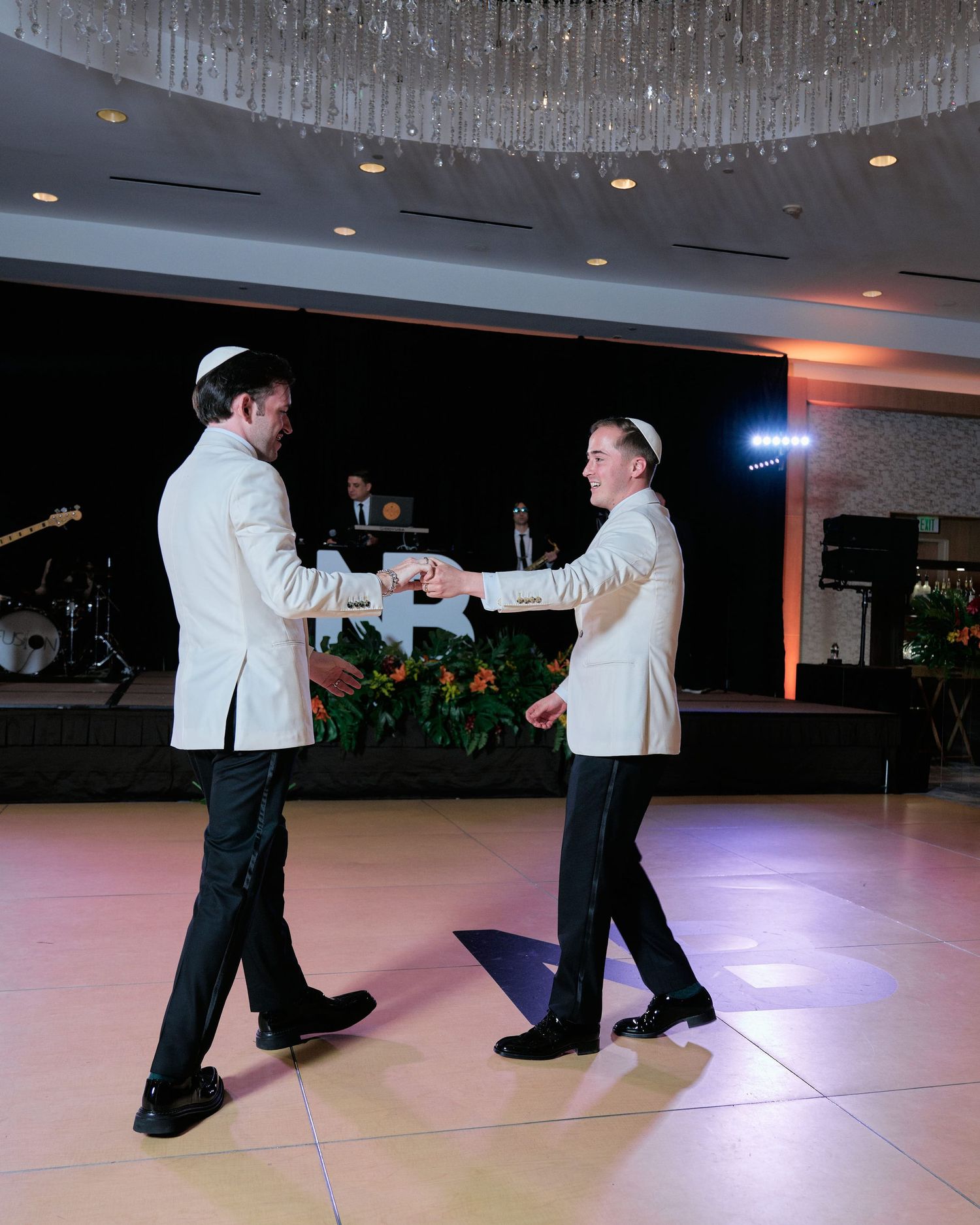 Two people wearing matching white jackets dance together on an illuminated ballroom floor with crystal chandeliers above.