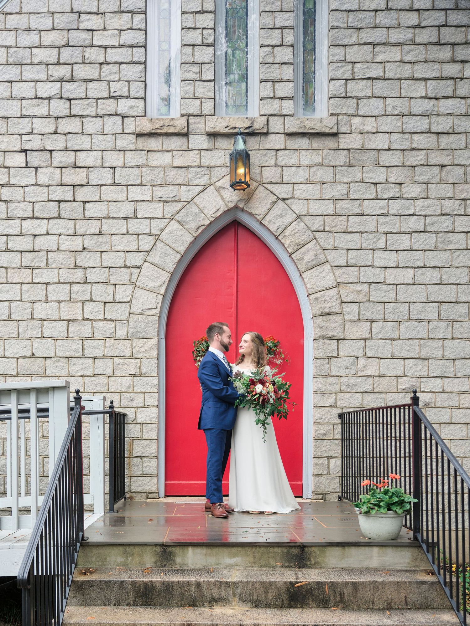 A couple stands on church steps against a bright red door and stone wall backdrop for their wedding photos.
