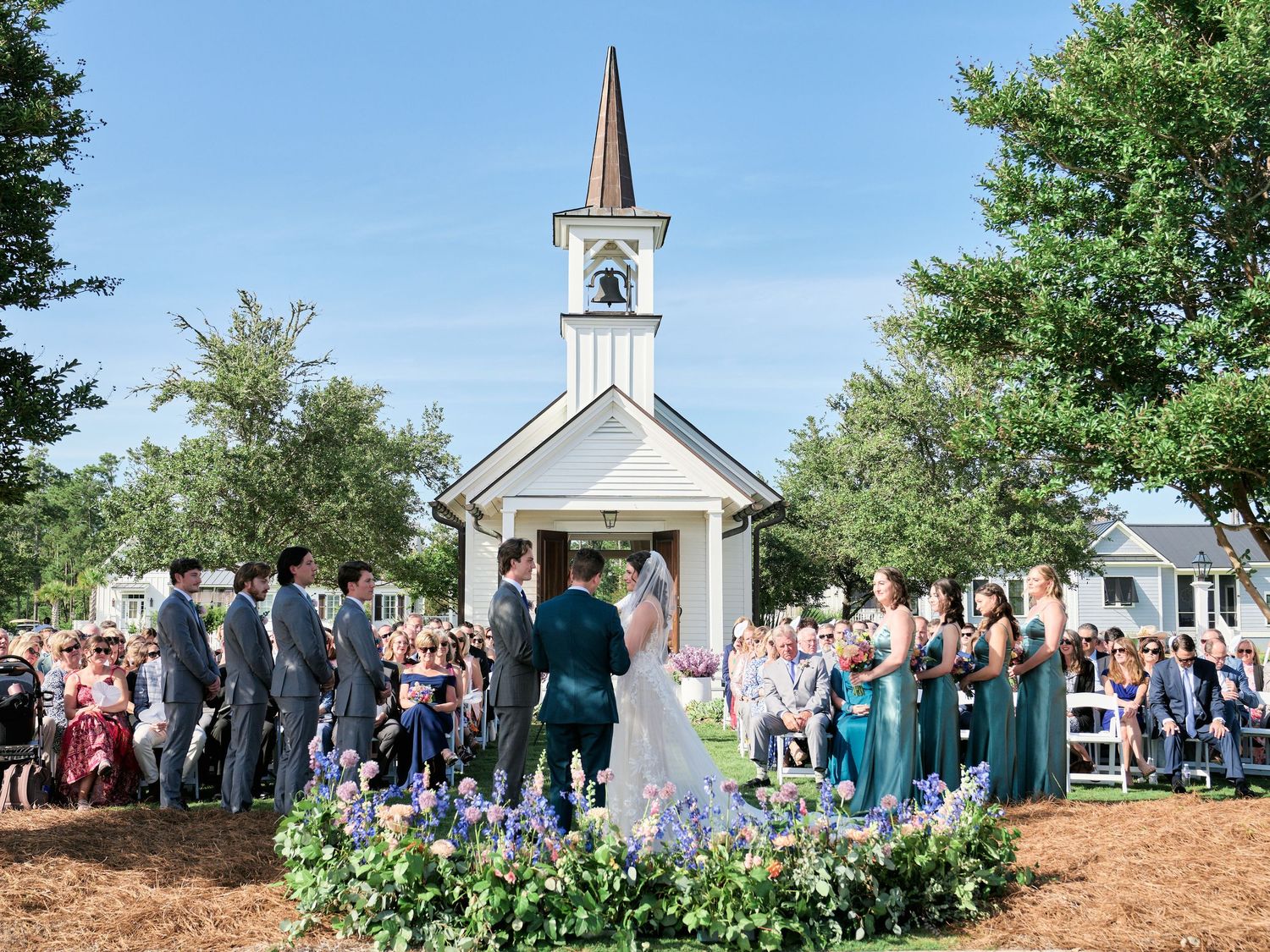 A small white chapel with a steeple sits among trees while a crowd gathers for an outdoor wedding ceremony.
