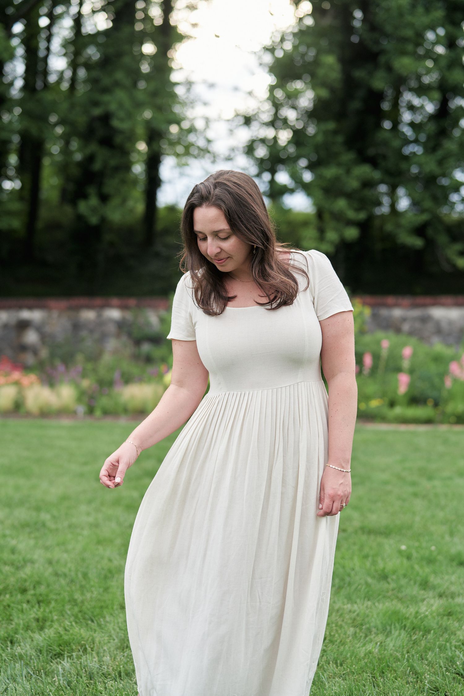 A person in a white maxi dress stands gracefully in a garden setting with trees in the background.