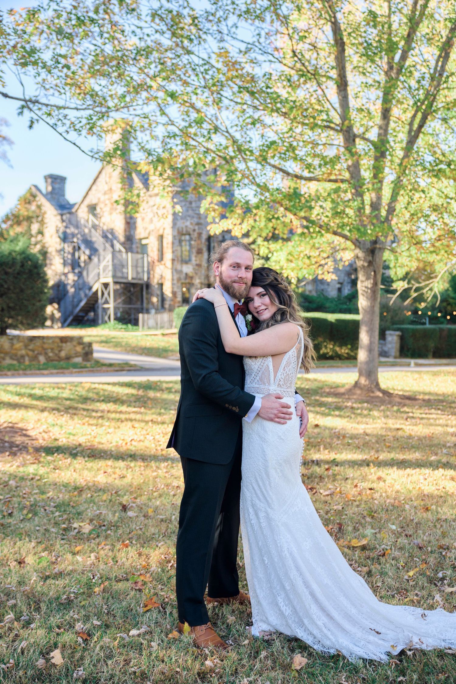 Bride and groom embrace on autumn lawn with historic stone building in background.
