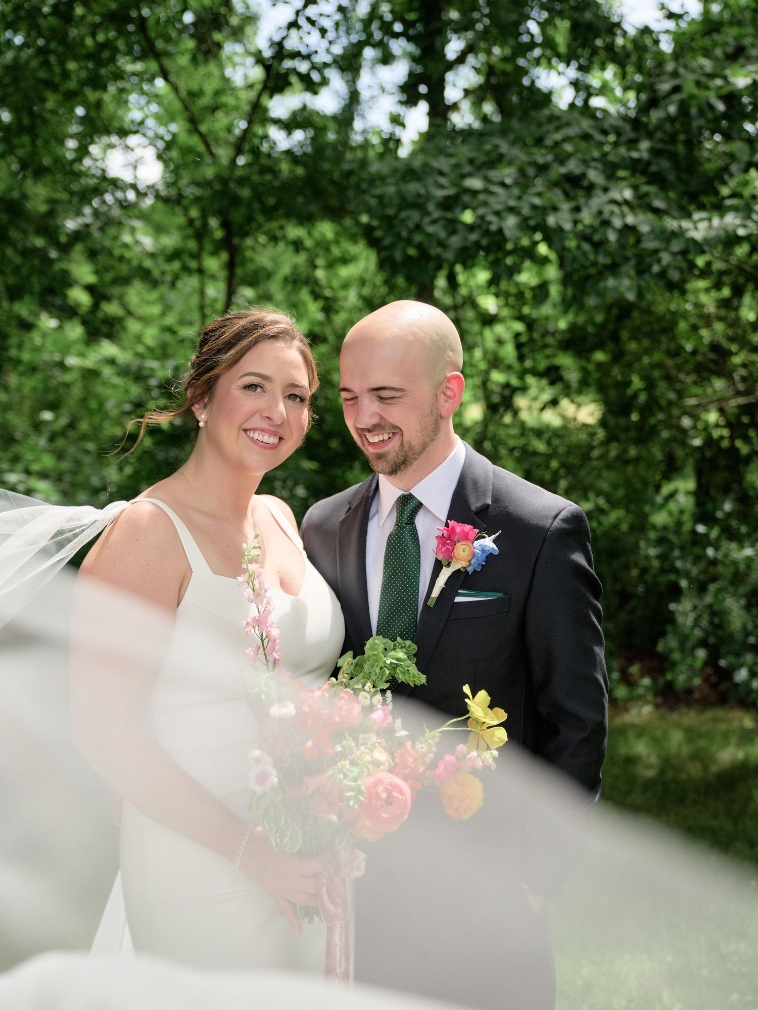 Intimate candid moment of couple smiling with bouquet and veil in foreground during wedding photos.