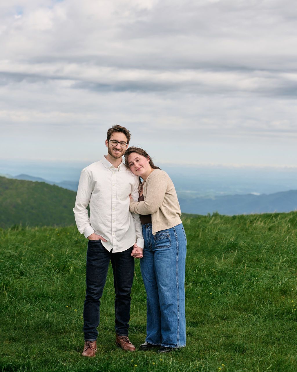 A couple stands together in a grassy mountain meadow under cloudy skies, dressed casually in light colored clothing.