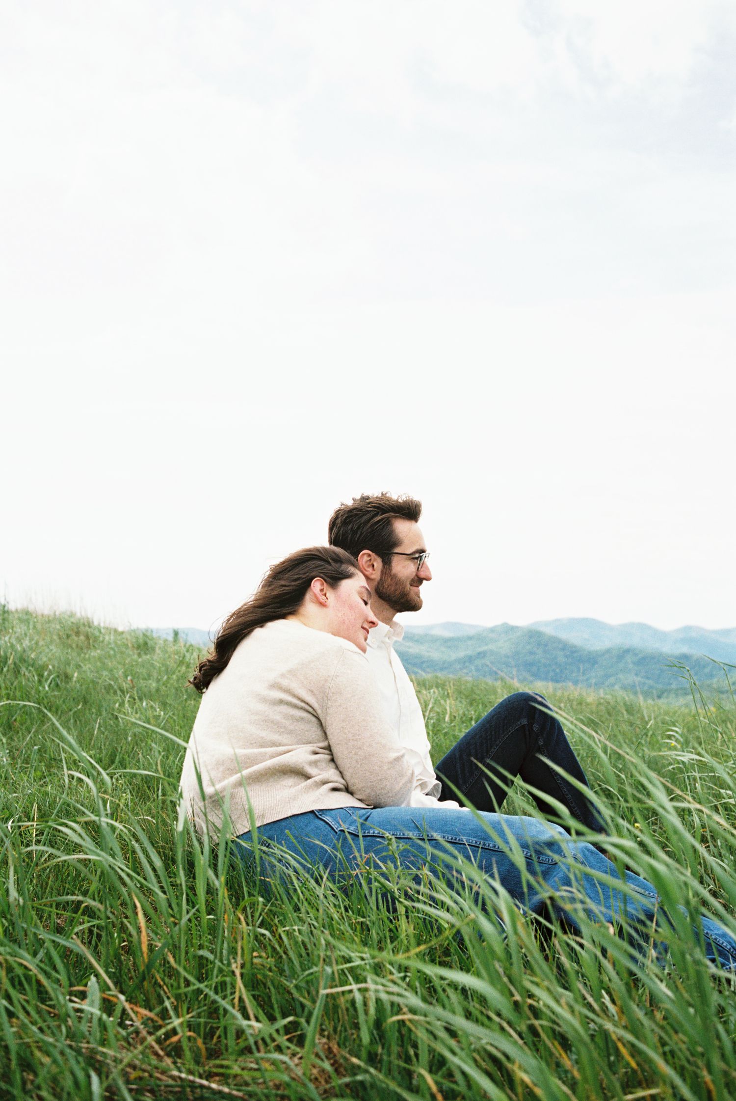 A couple sits together in tall grass on a hillside overlooking mountains on a peaceful afternoon.