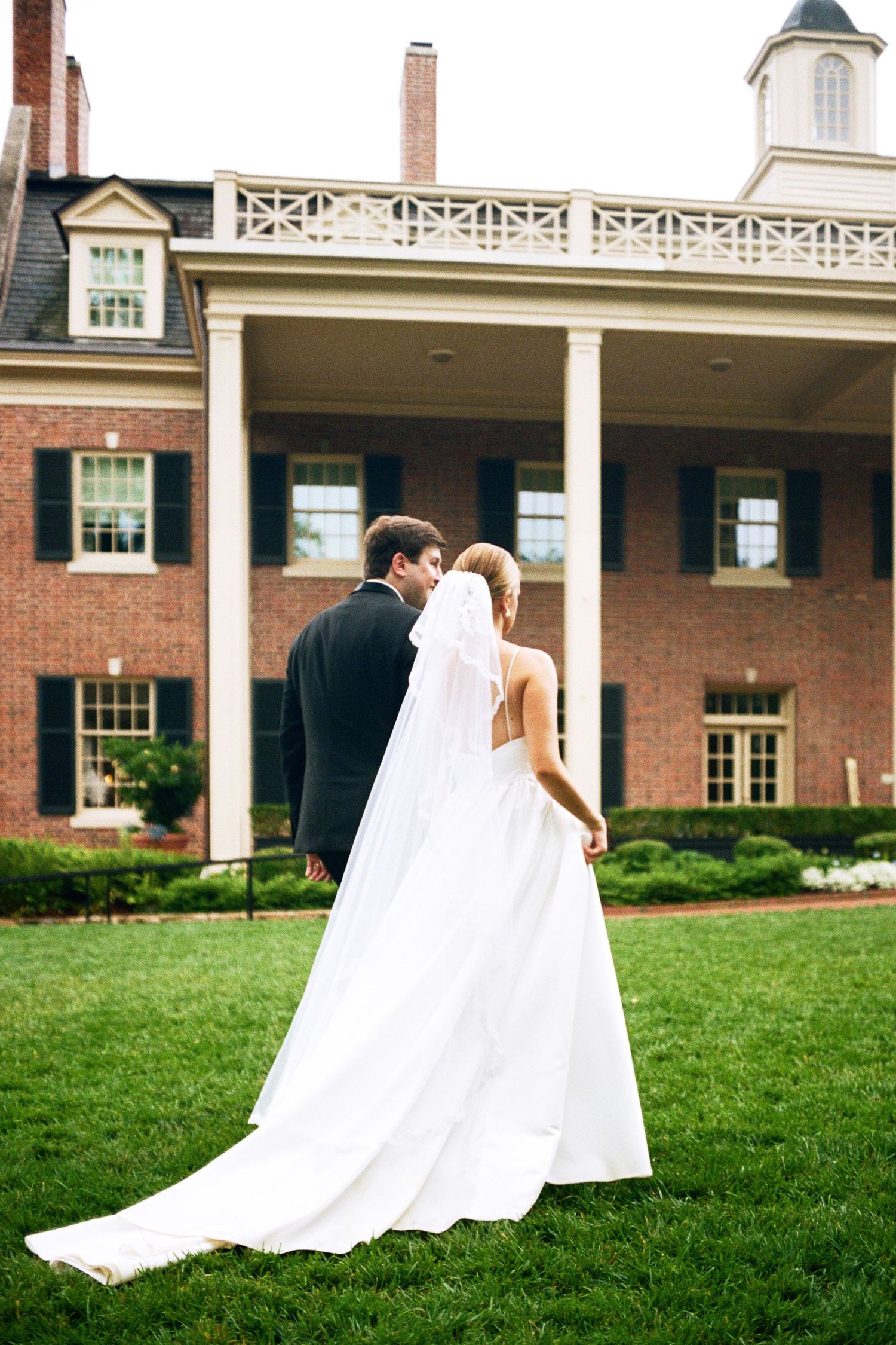 Bride in flowing white gown and groom embrace on the lawn of a beautiful brick mansion.