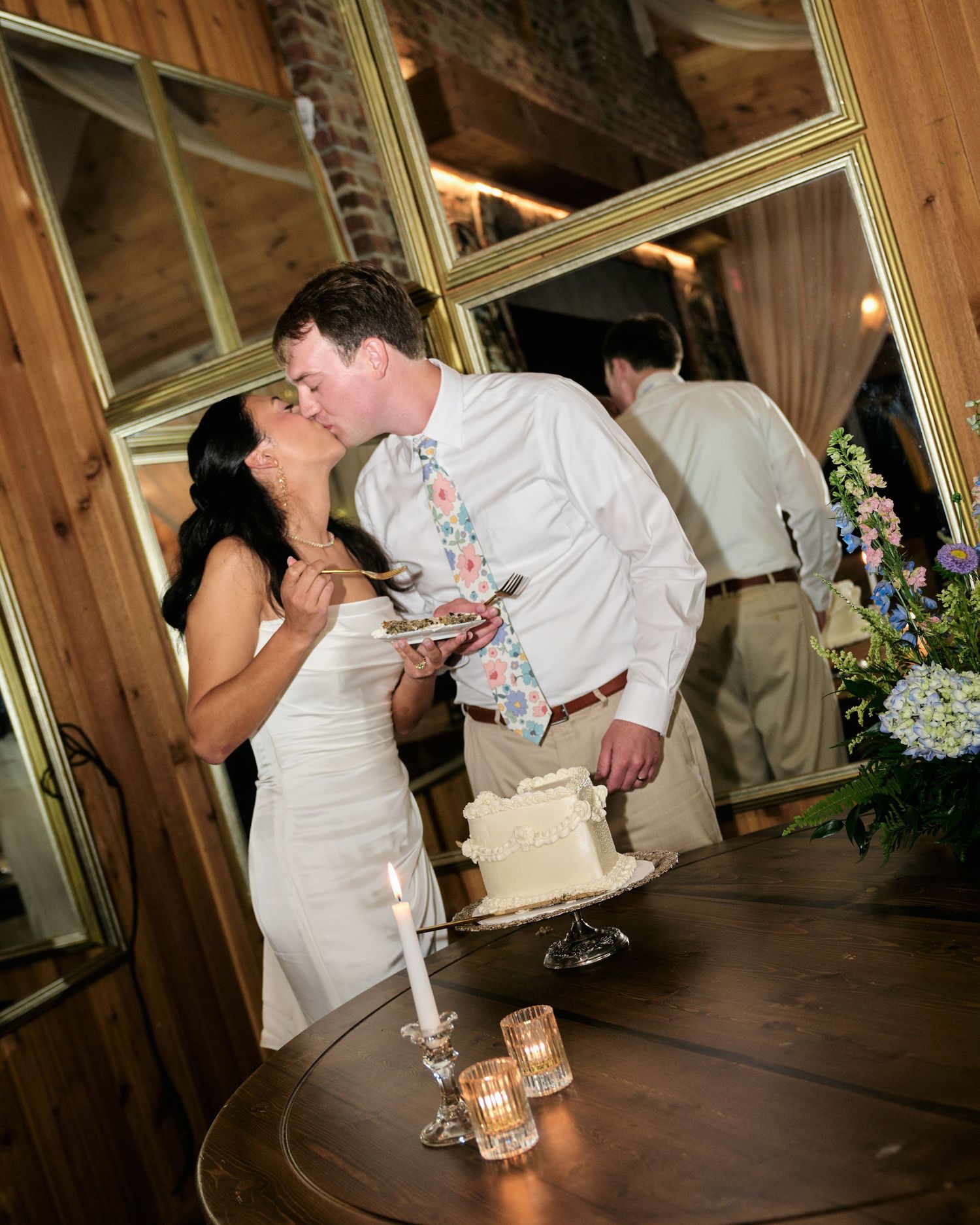 Bride and groom share a kiss while cutting their white wedding cake at a rustic indoor reception venue.