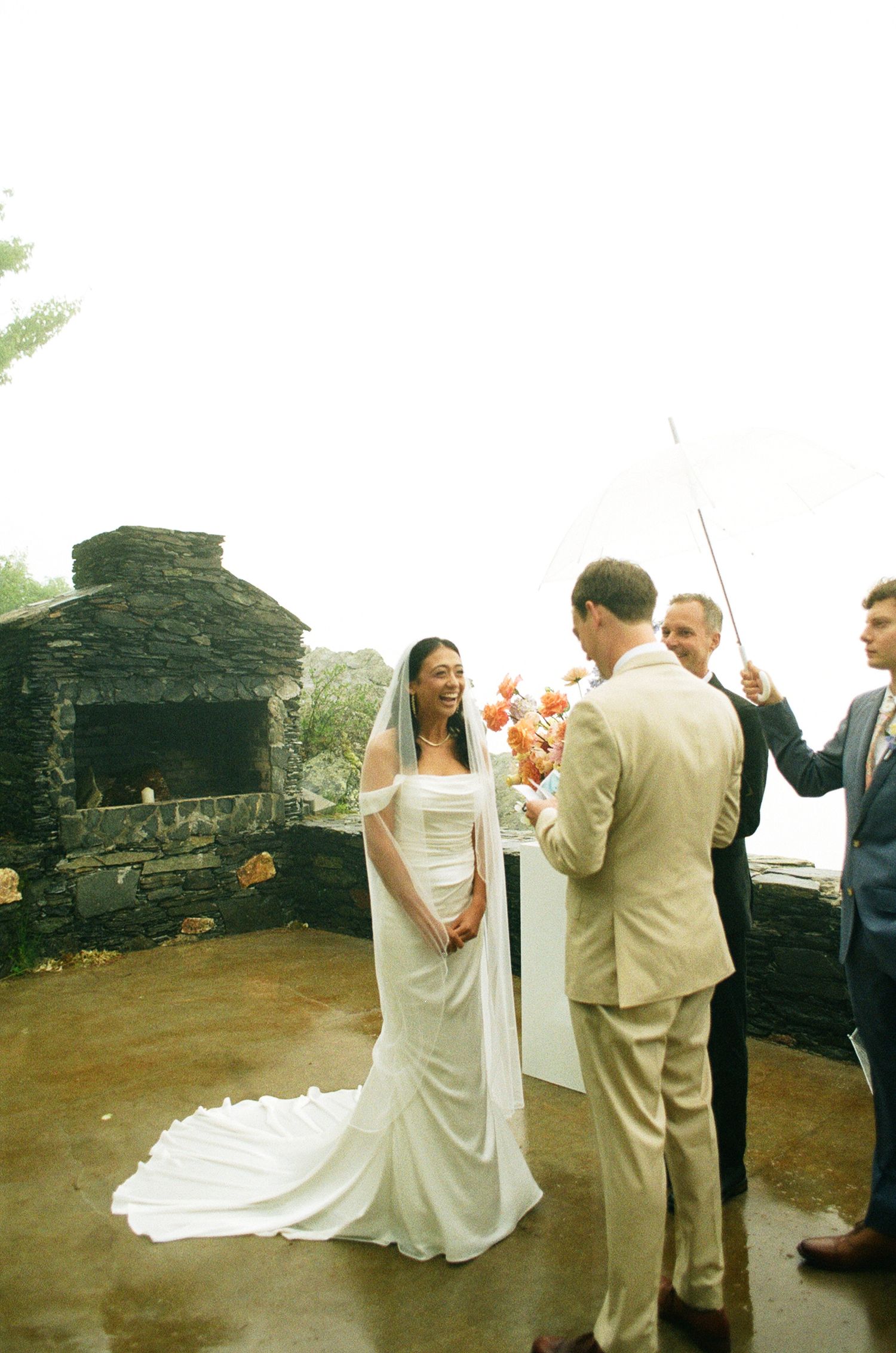 Bride in white gown with veil stands at outdoor stone fireplace altar with groom in tan suit during wedding ceremony.