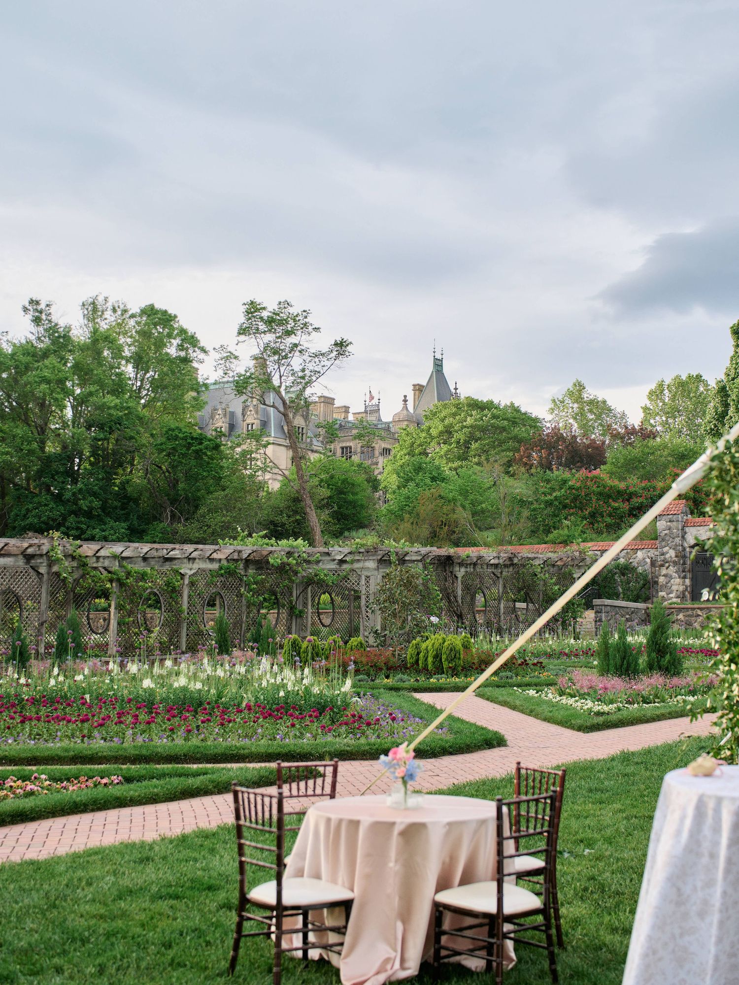Elegant garden wedding setup with white tablecloths and wooden chairs along a curved brick pathway surrounded by blooming flowers.