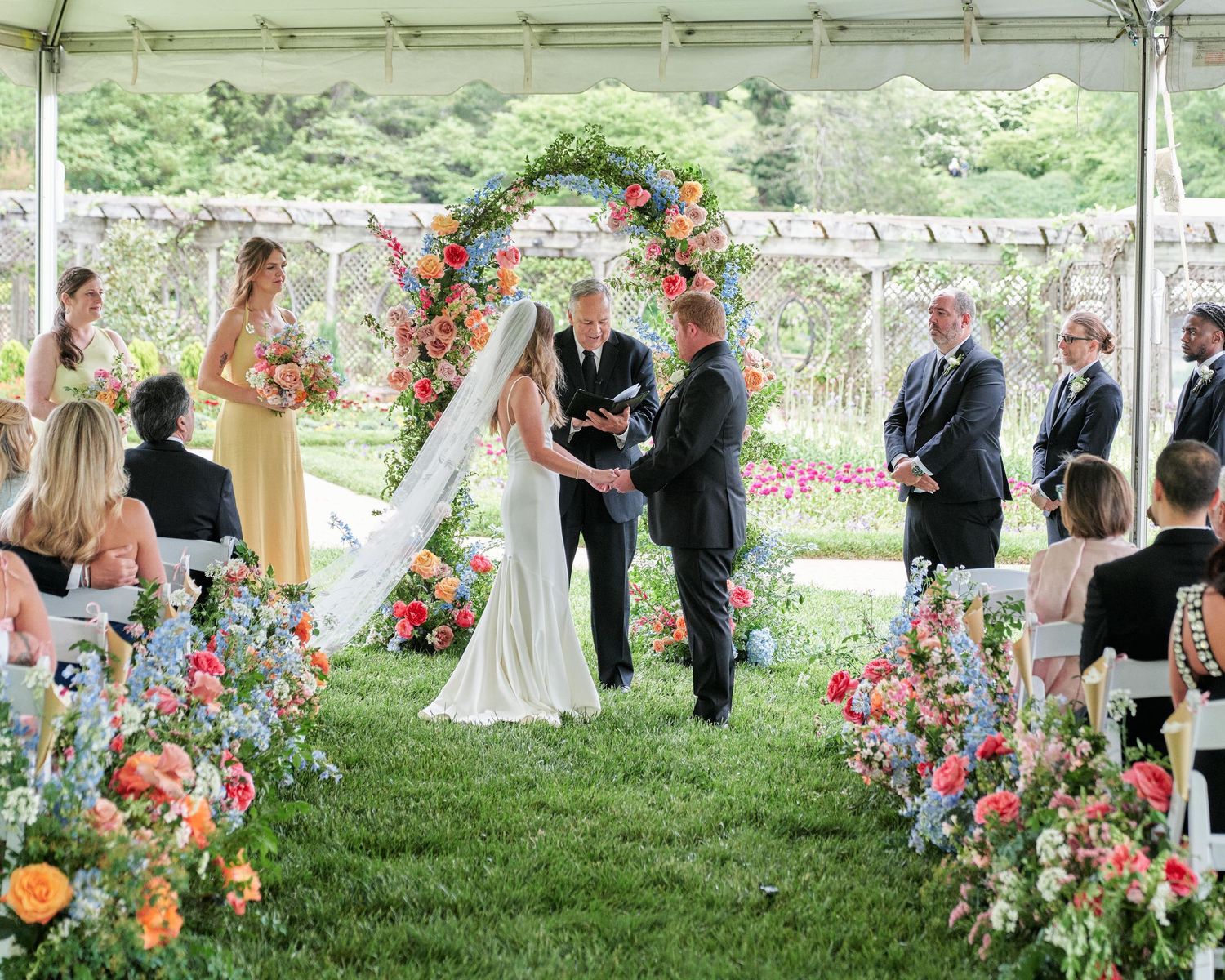 Outdoor wedding ceremony under white tent with colorful floral arch and aisle decorations on lush green lawn.