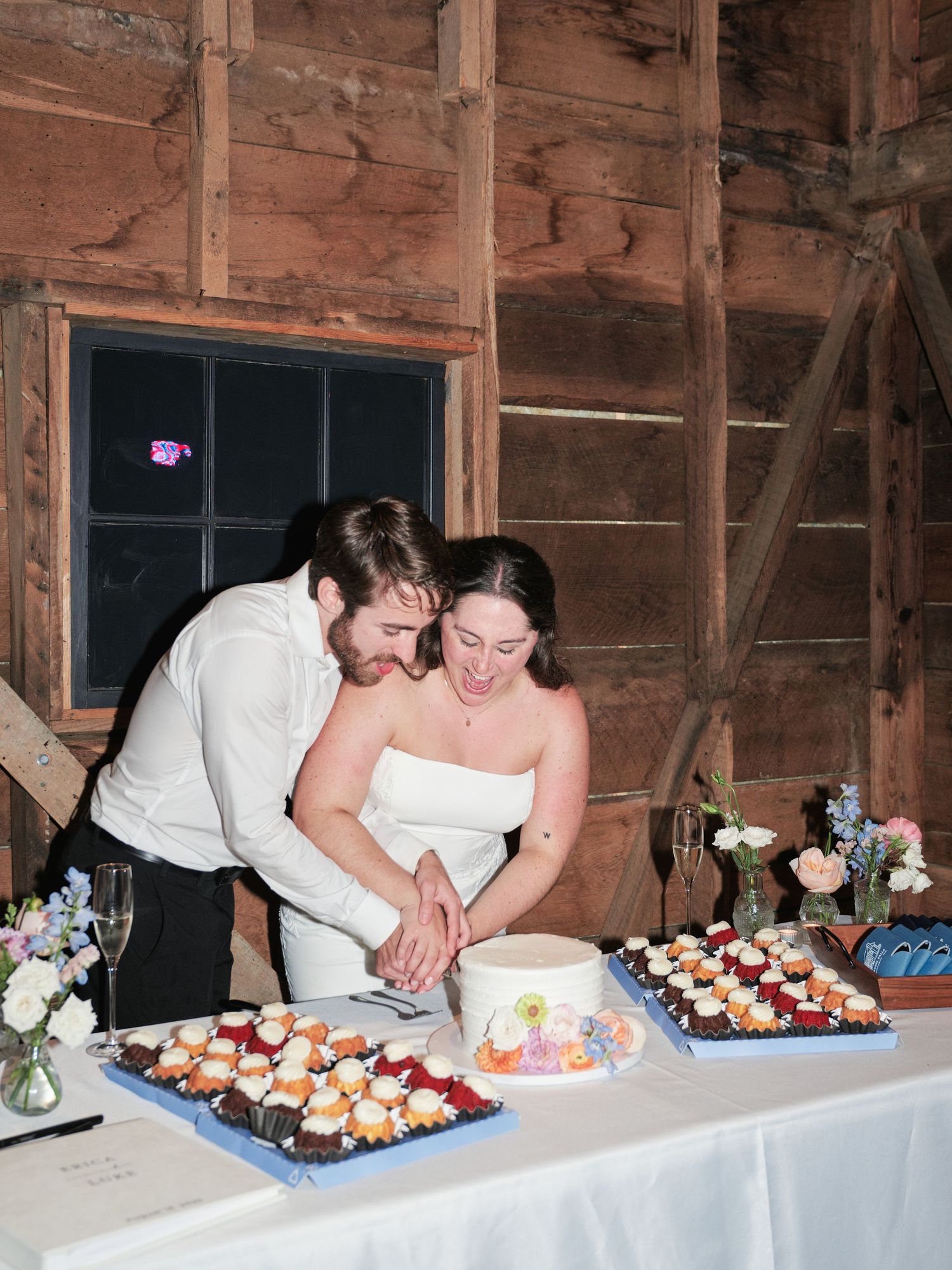 Bride and groom cutting wedding cake together in a rustic barn venue with dessert trays on white tablecloth.