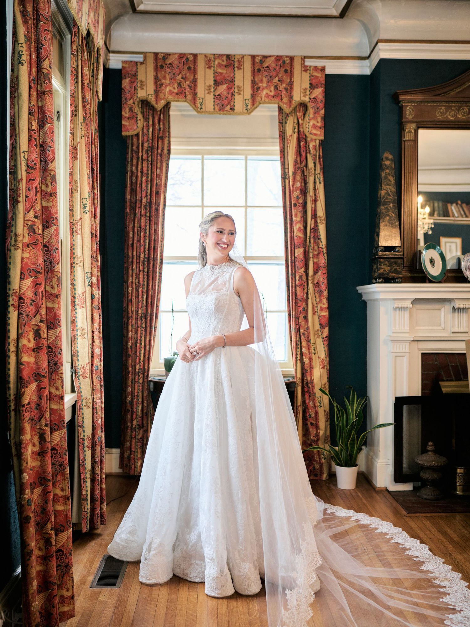 Bride in elegant white ball gown with lace train stands by window in classic room with floral curtains and fireplace.