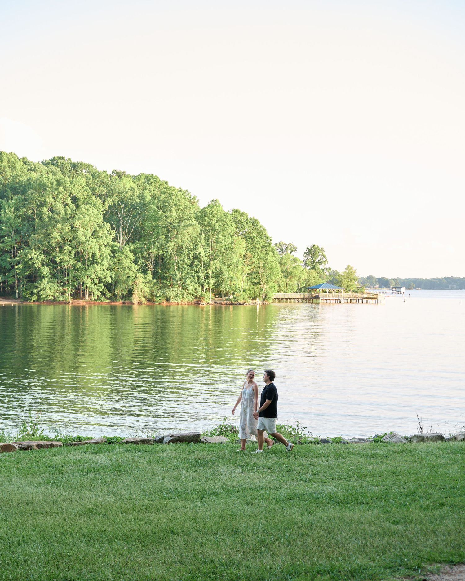 Couple walking on a serene lakeside scene with lush green trees and grass along the shoreline during a summer day.