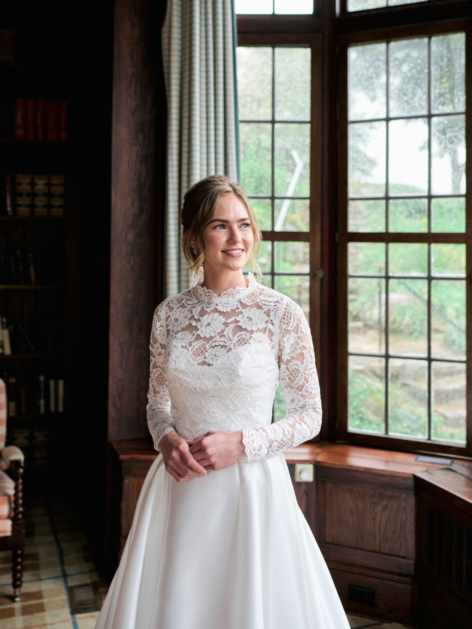 Bride in elegant long-sleeve lace wedding gown standing by a window in a wood-paneled room, smiling softly.
