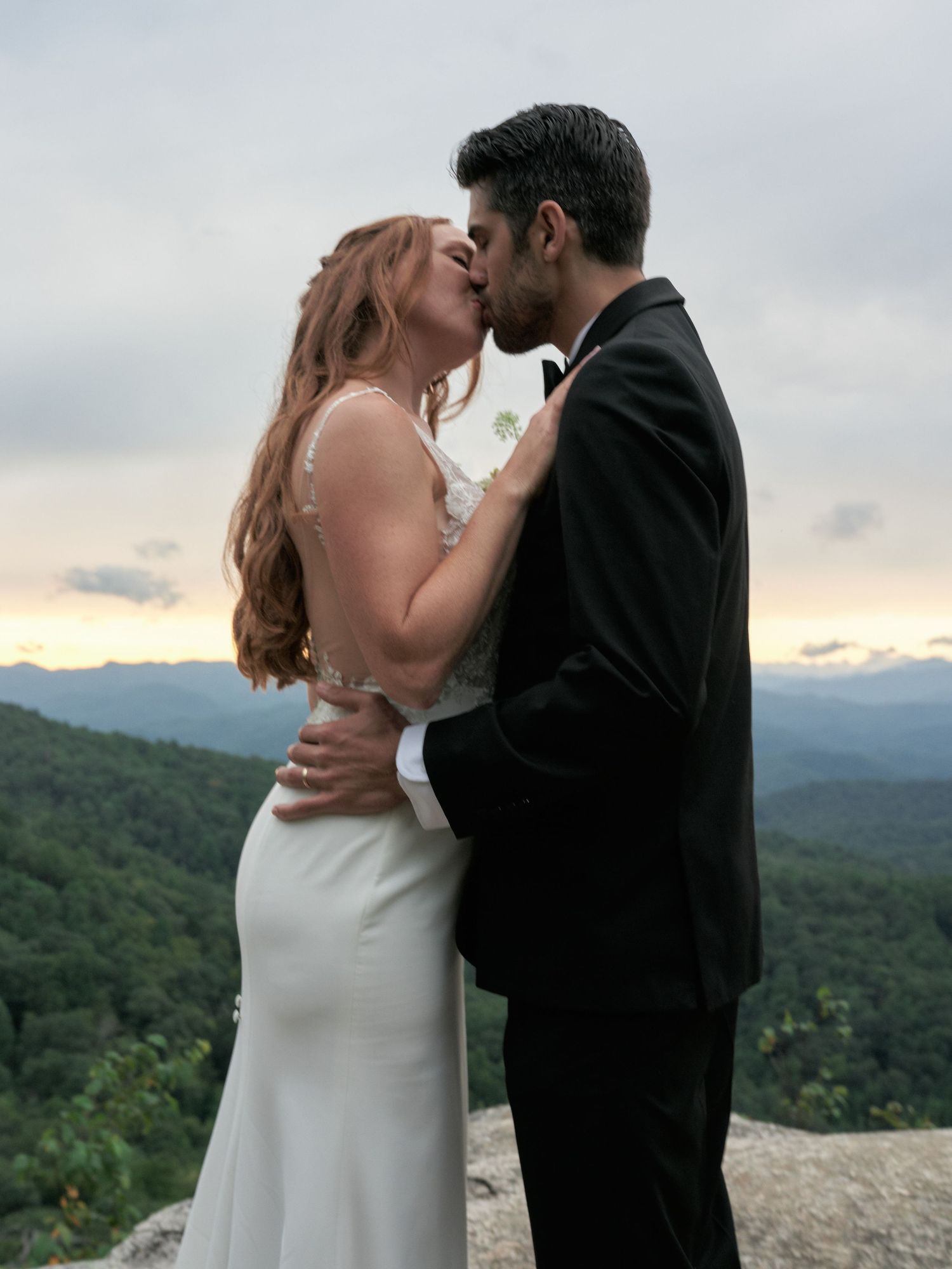 A romantic wedding moment captured on a mountain cliff with scenic rolling hills in the background at dusk.