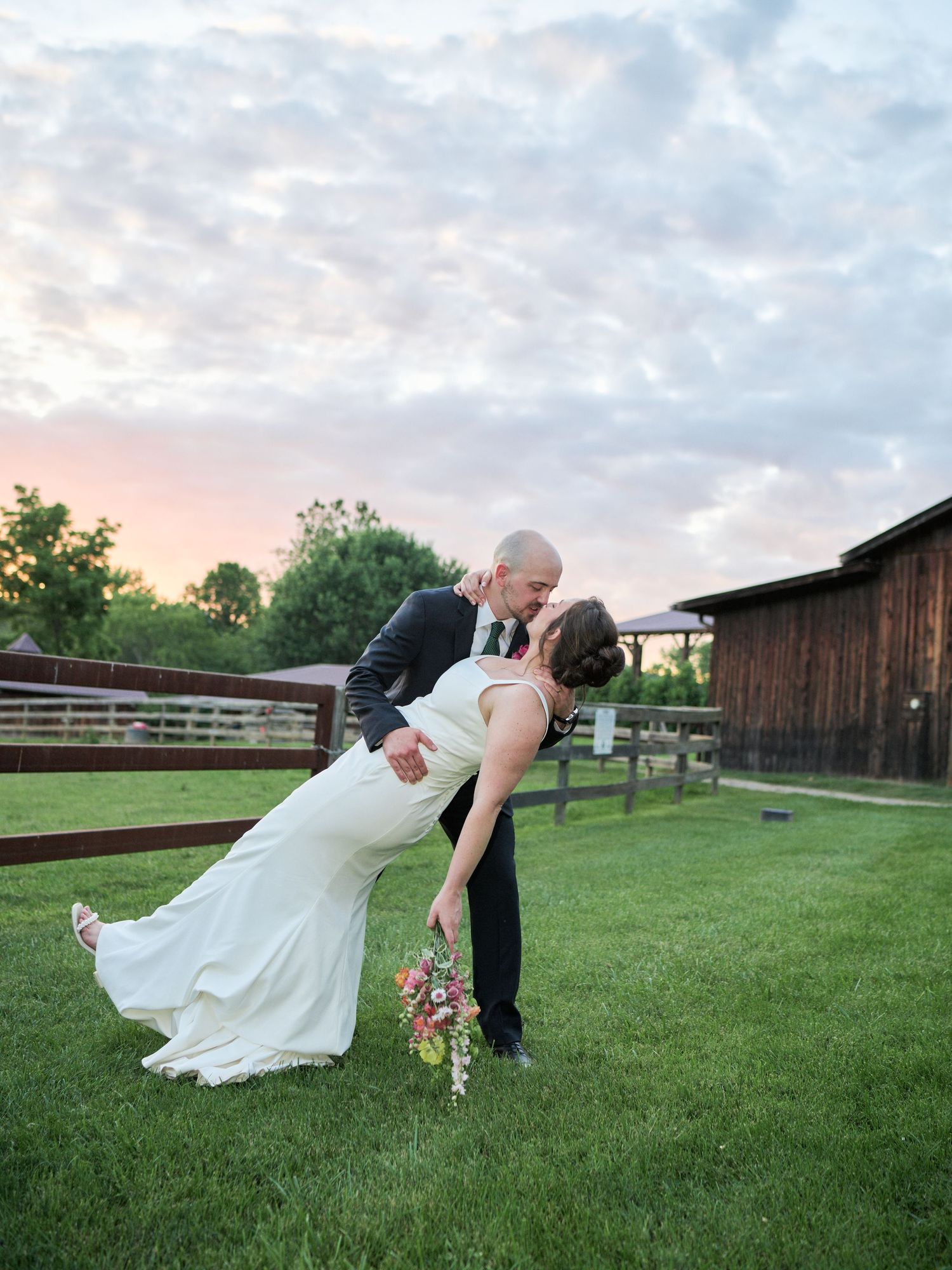 A romantic dip pose during wedding photos on a grassy lawn with a barn in the background.