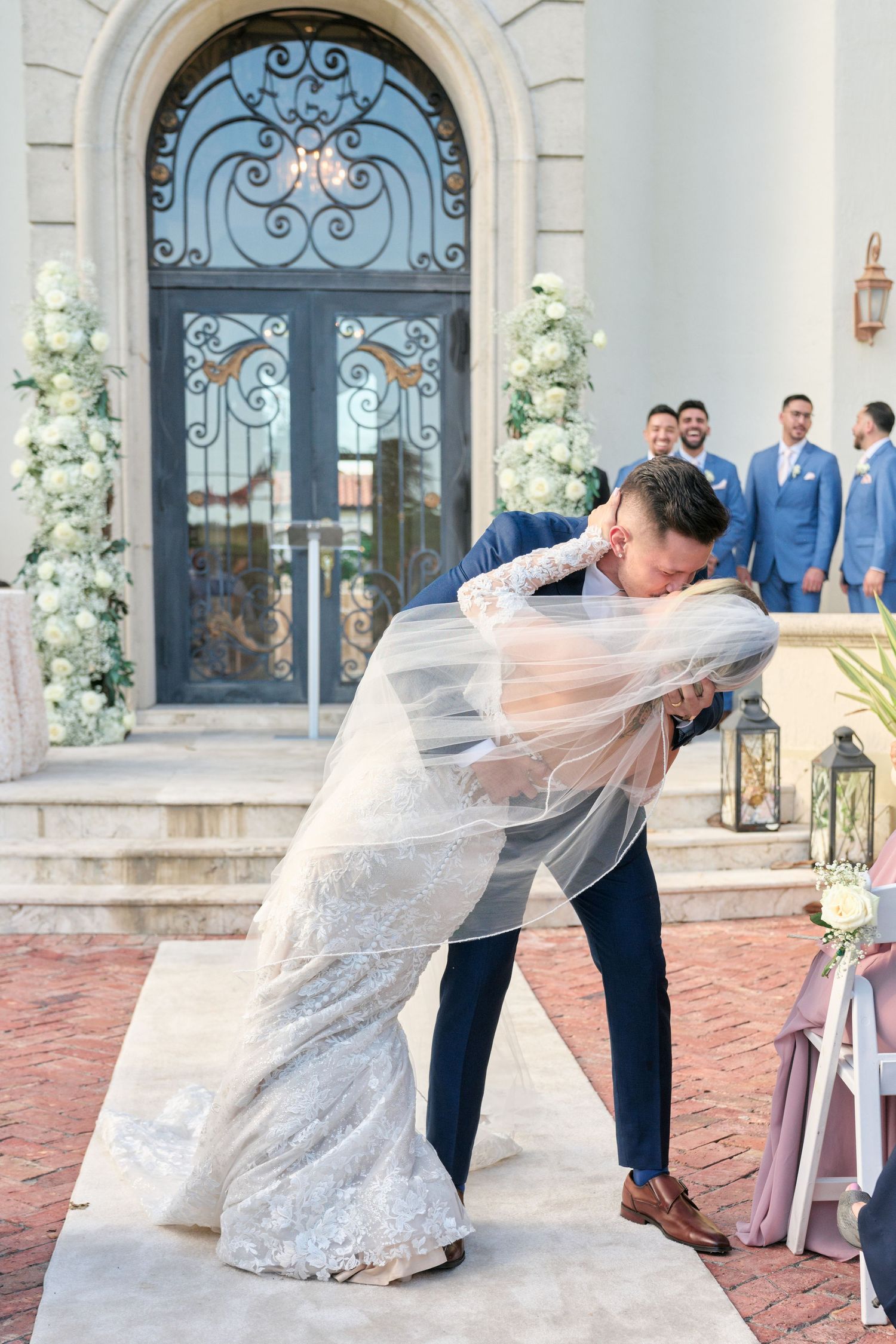 Dramatic dip kiss between newlyweds in front of ornate blue door with floral decor.