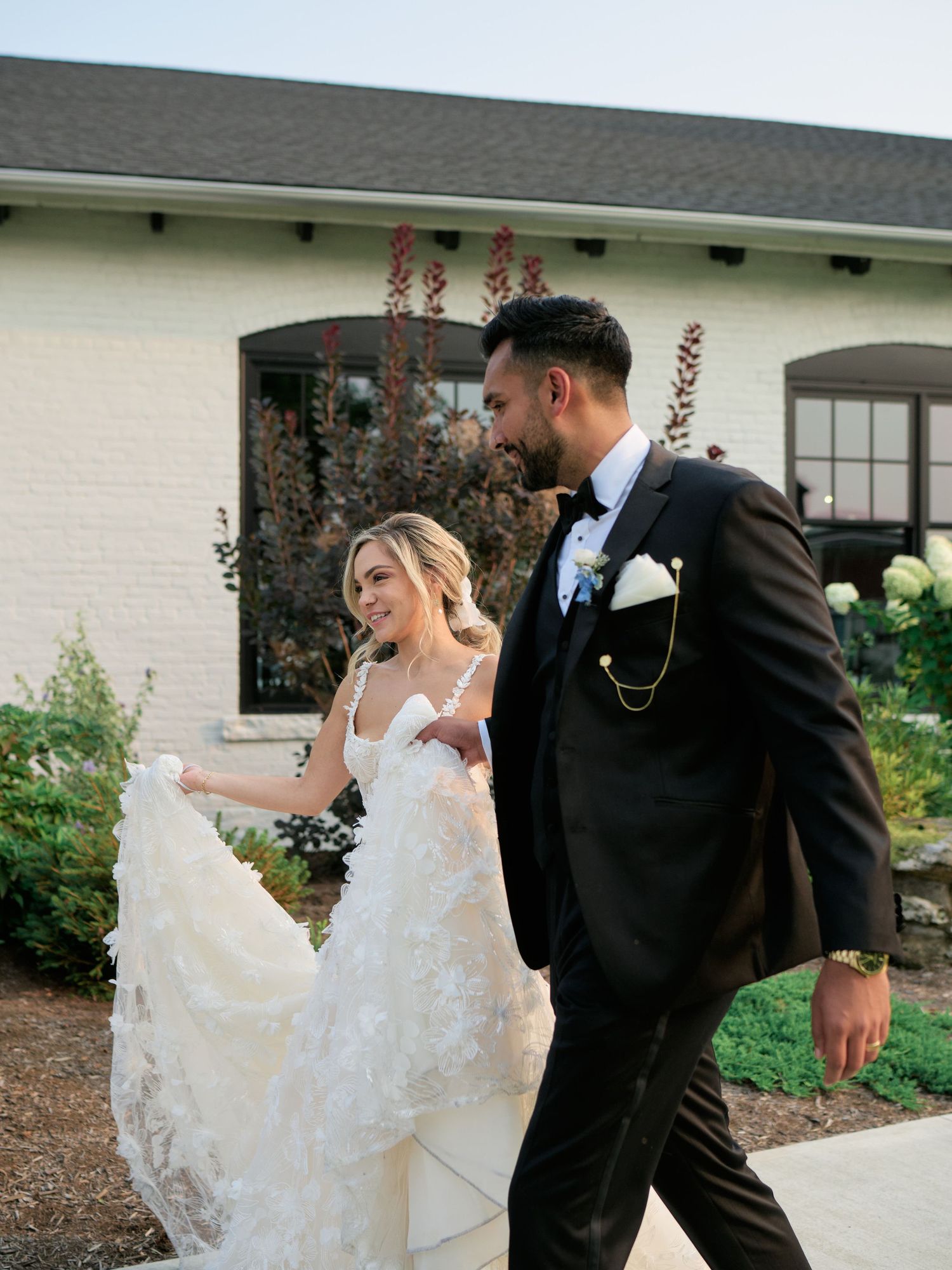 A couple walking together outdoors, with the bride in a flowing white lace dress and the groom in a classic black suit.