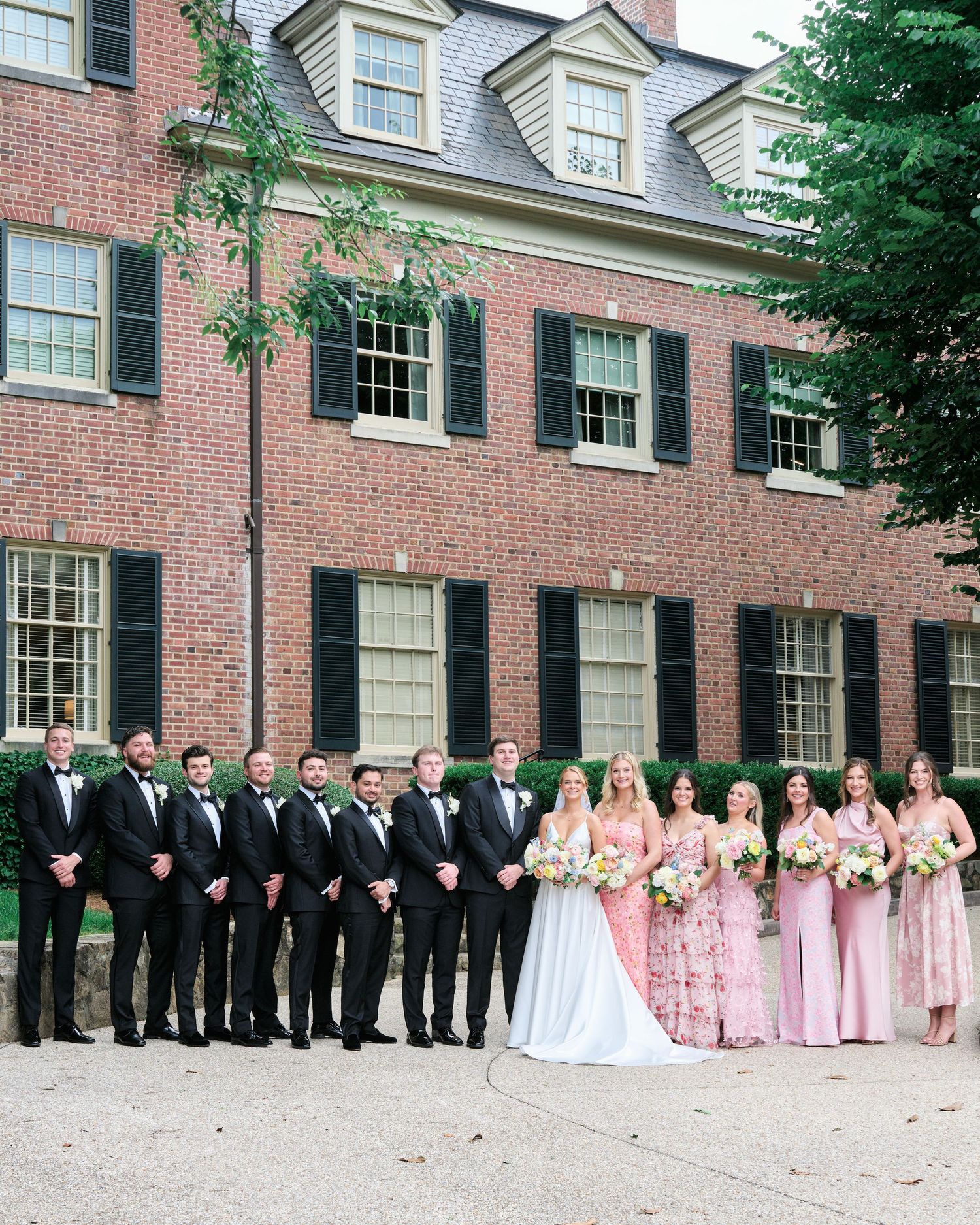 Full wedding party lined up for formal photo outside historic brick mansion with black shutters.