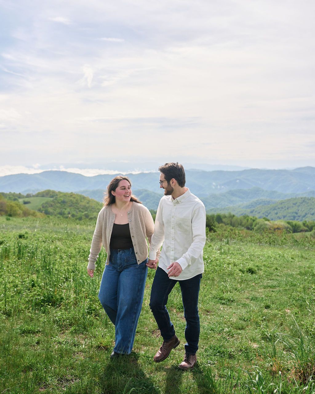 A couple walks together on a grassy hilltop with scenic mountain views in the background on a sunny day.