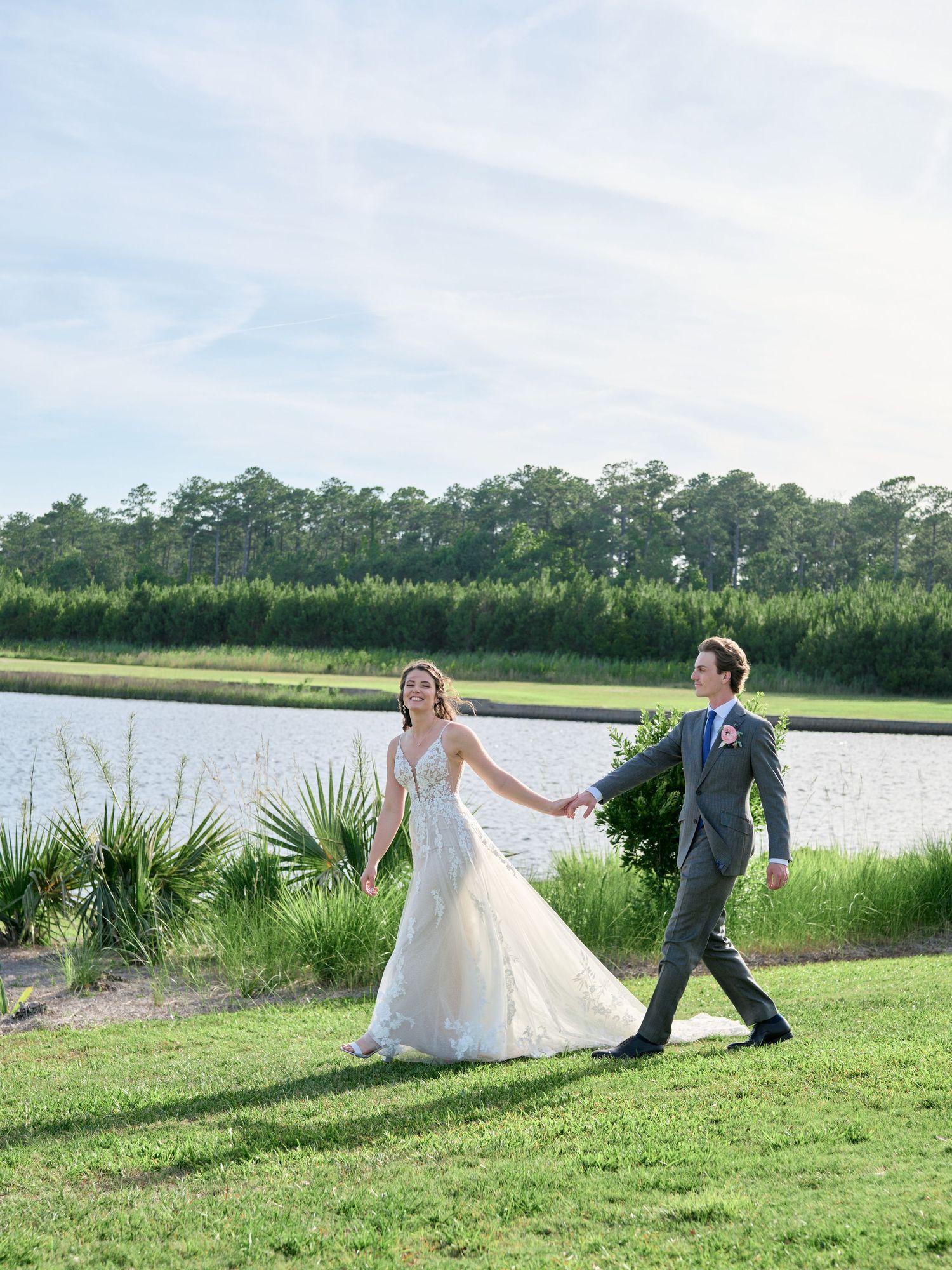 A couple walks hand in hand along a grassy waterfront path with tall pine trees in the background on their wedding day.
