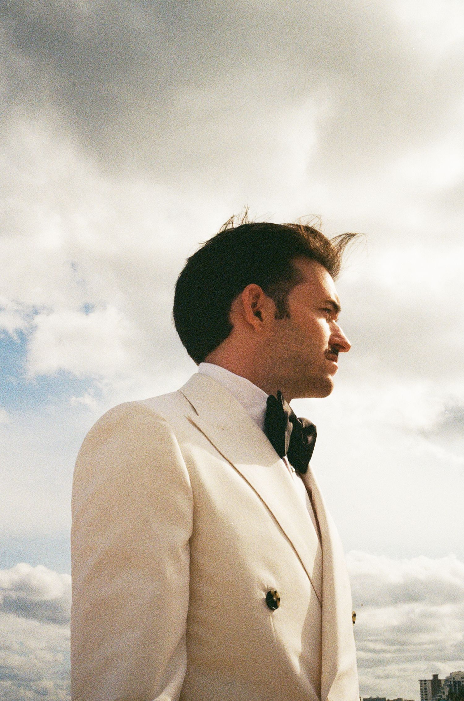 Profile view of person in cream double-breasted jacket against cloudy sky background.