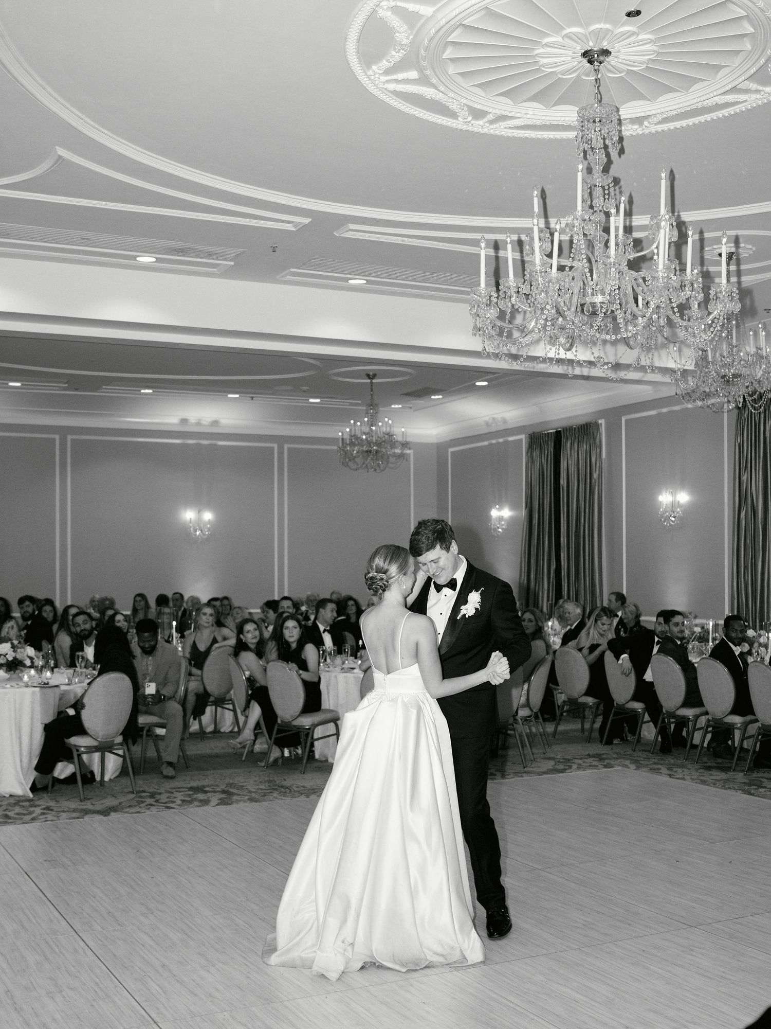 A couple shares their first dance under a crystal chandelier in an elegant ballroom.