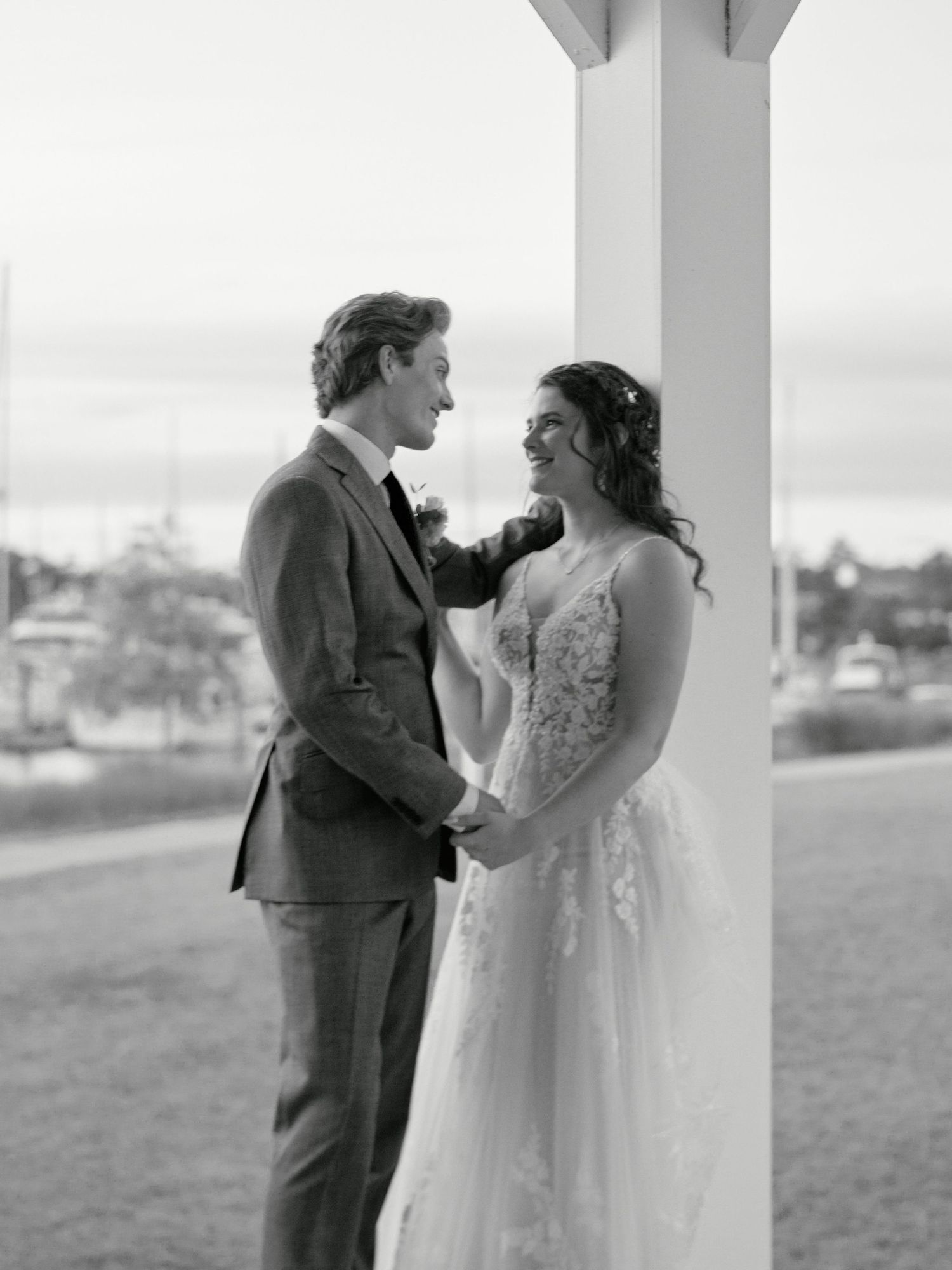A romantic black and white wedding photo of a couple sharing an intimate moment beside a column with a waterfront view.