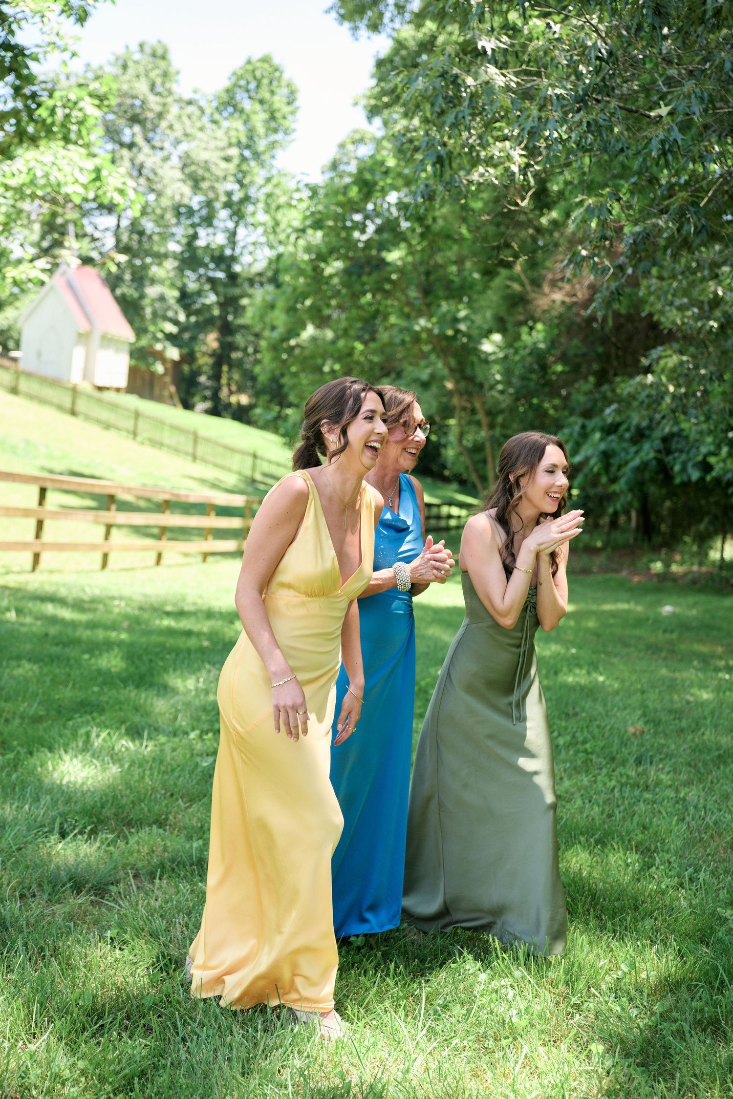 Three bridesmaids in colorful dresses laughing together on a sunny lawn.