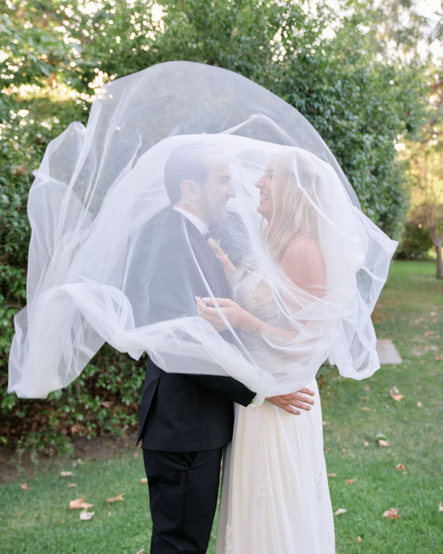 Creative wedding photo with veil flowing over couple in garden setting.