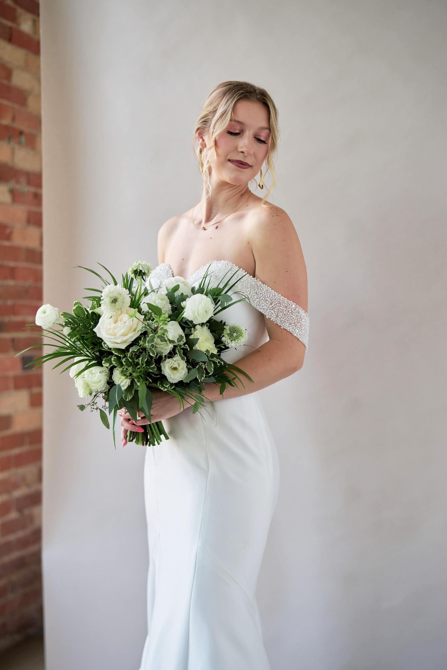 Elegant bride in off-shoulder white gown holds a luxurious bouquet of white roses and greenery against brick wall.