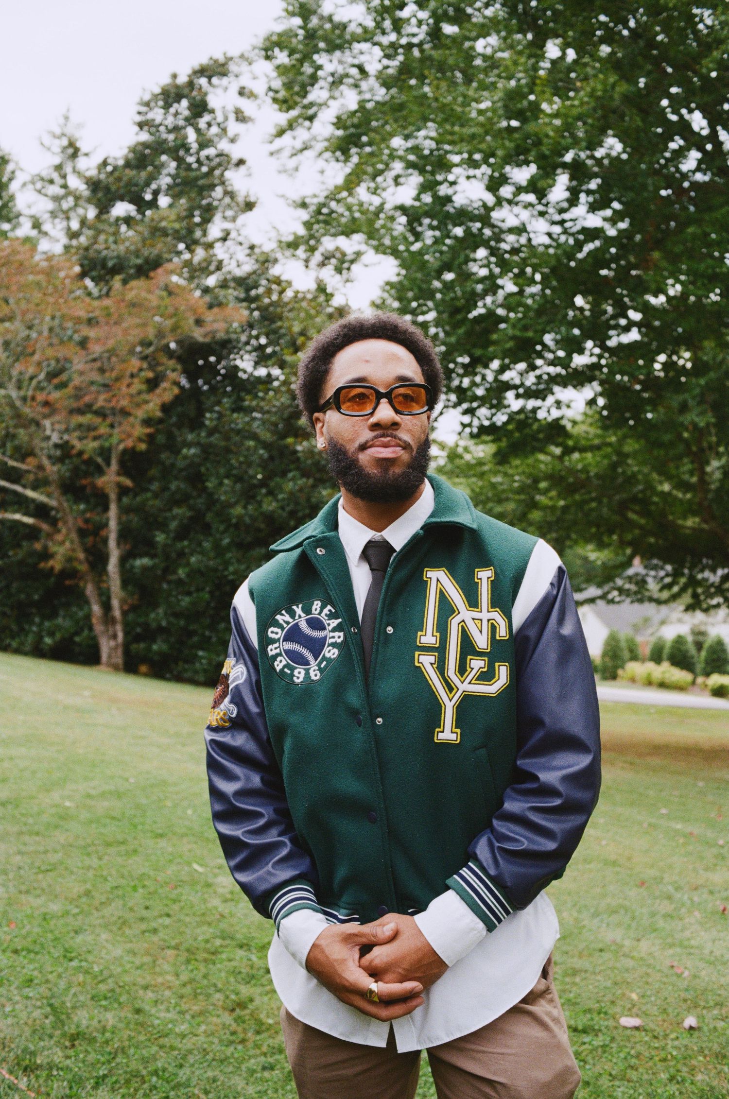 Man wearing vintage green and navy varsity jacket stands outdoors on grassy lawn.