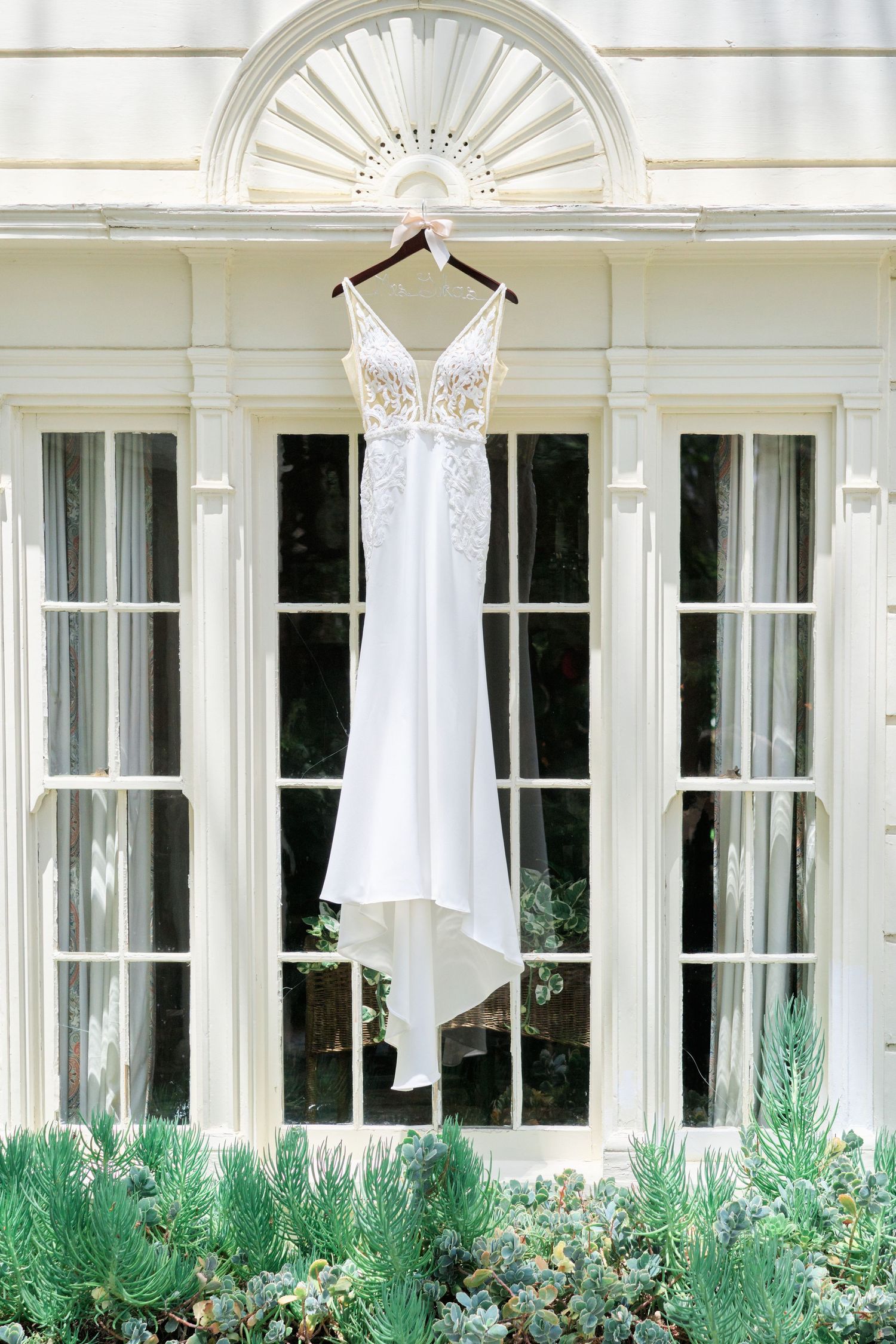 An elegant white wedding dress hangs against a classic white facade with fan window detail.