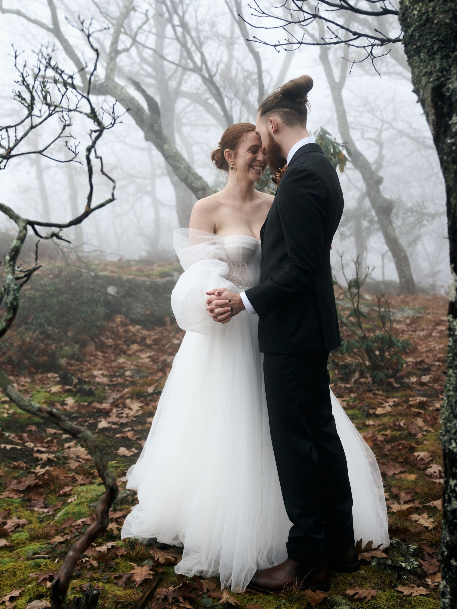 A romantic wedding couple embraces in a misty forest setting with bare trees and fallen leaves.