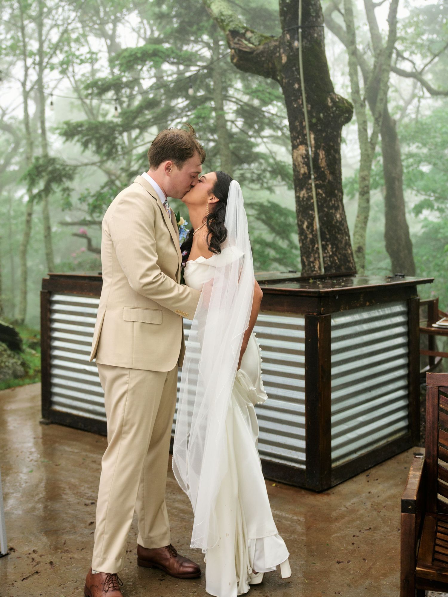 Bride and groom share first kiss in white wedding dress and tan suit outdoors in misty forest setting.