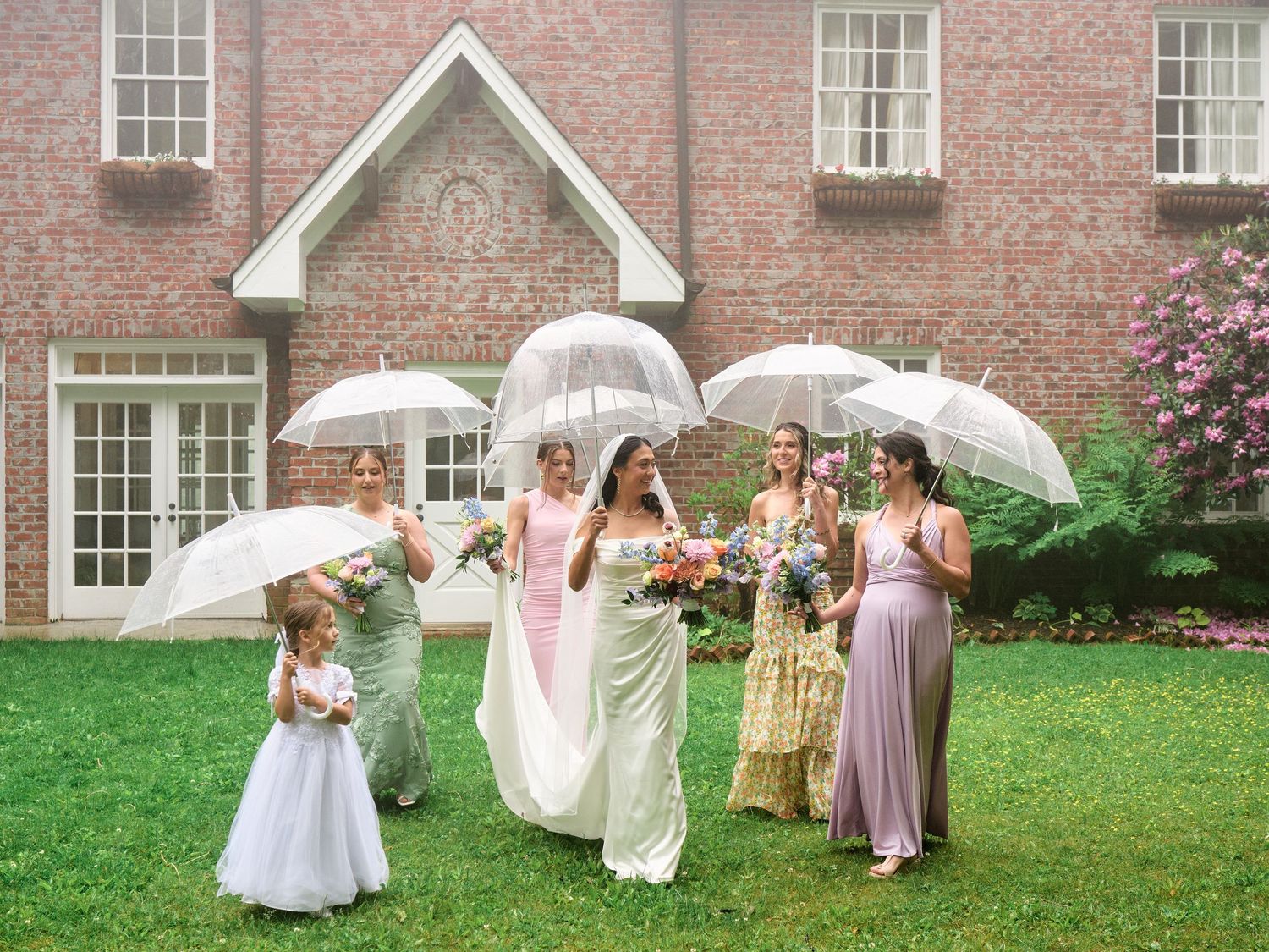 Bride and bridesmaids in pastel gowns holding white parasols walk across green lawn beside brick manor house.