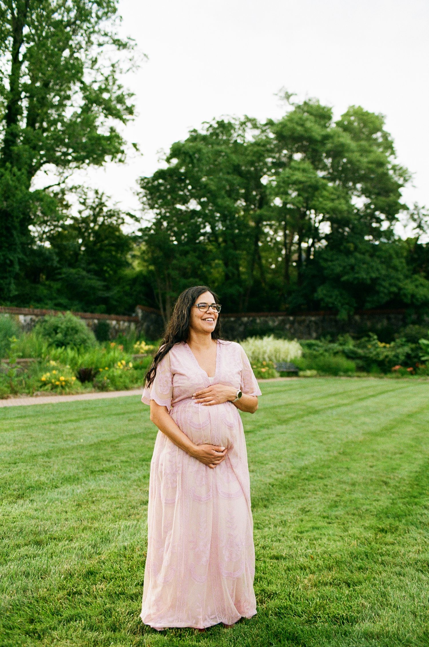 Pregnant woman in pink maternity dress holding belly, standing on green lawn with garden background.