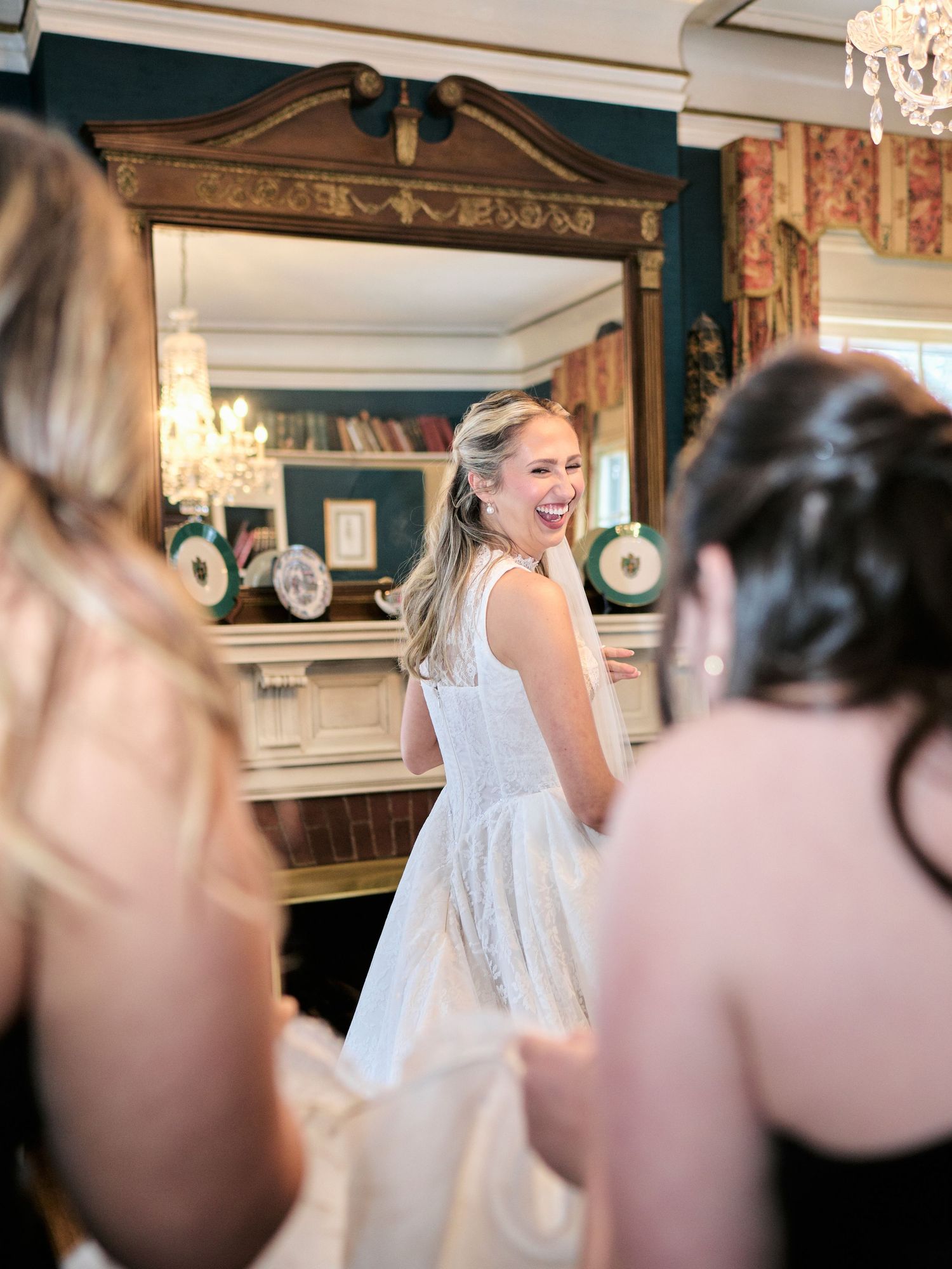 Bride in white wedding gown laughing joyfully with bridesmaids in elegant room with ornate mirror.