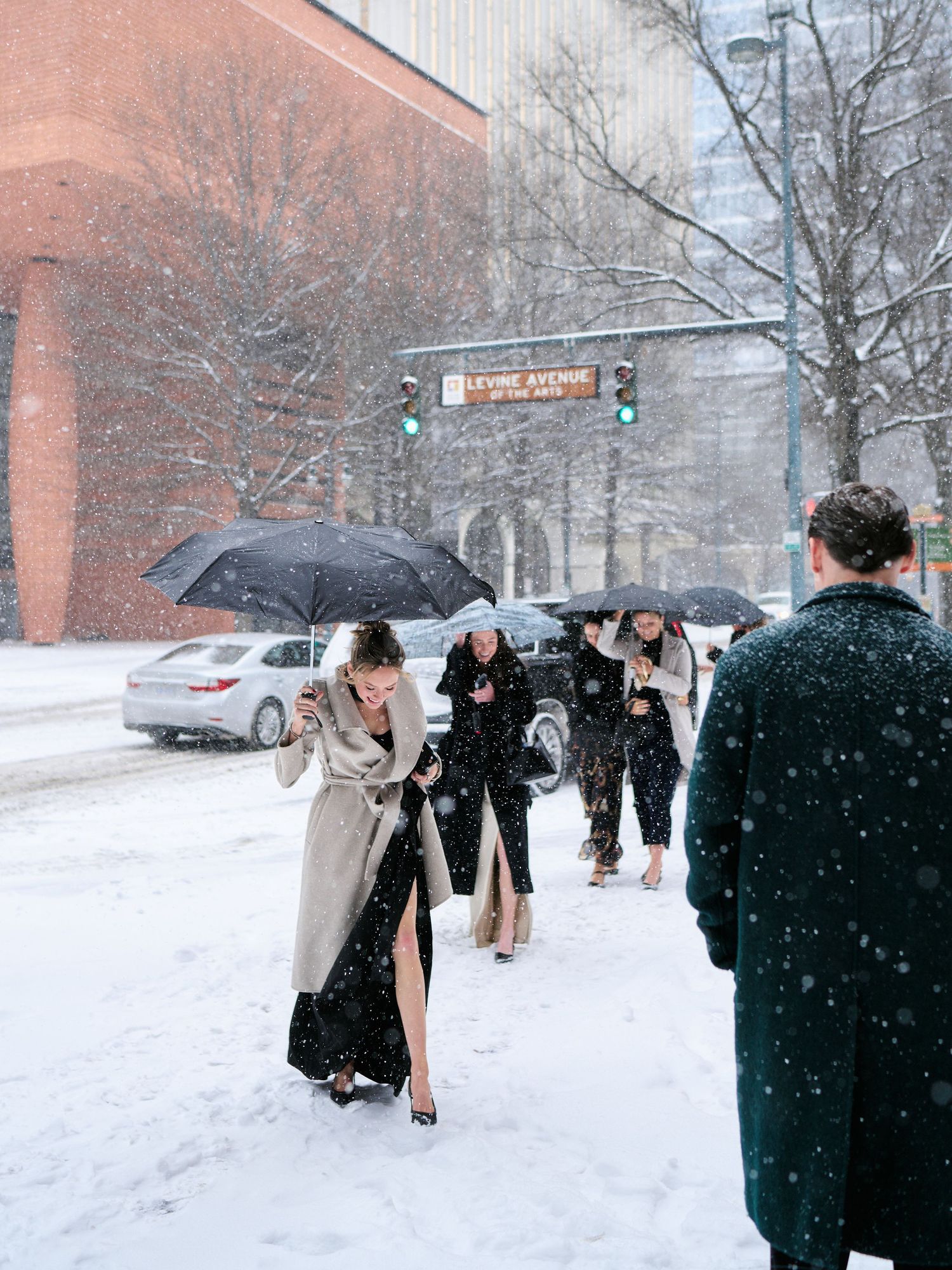 Elegantly dressed women walking in heavy snowfall on a city street, carrying black umbrellas in winter attire.