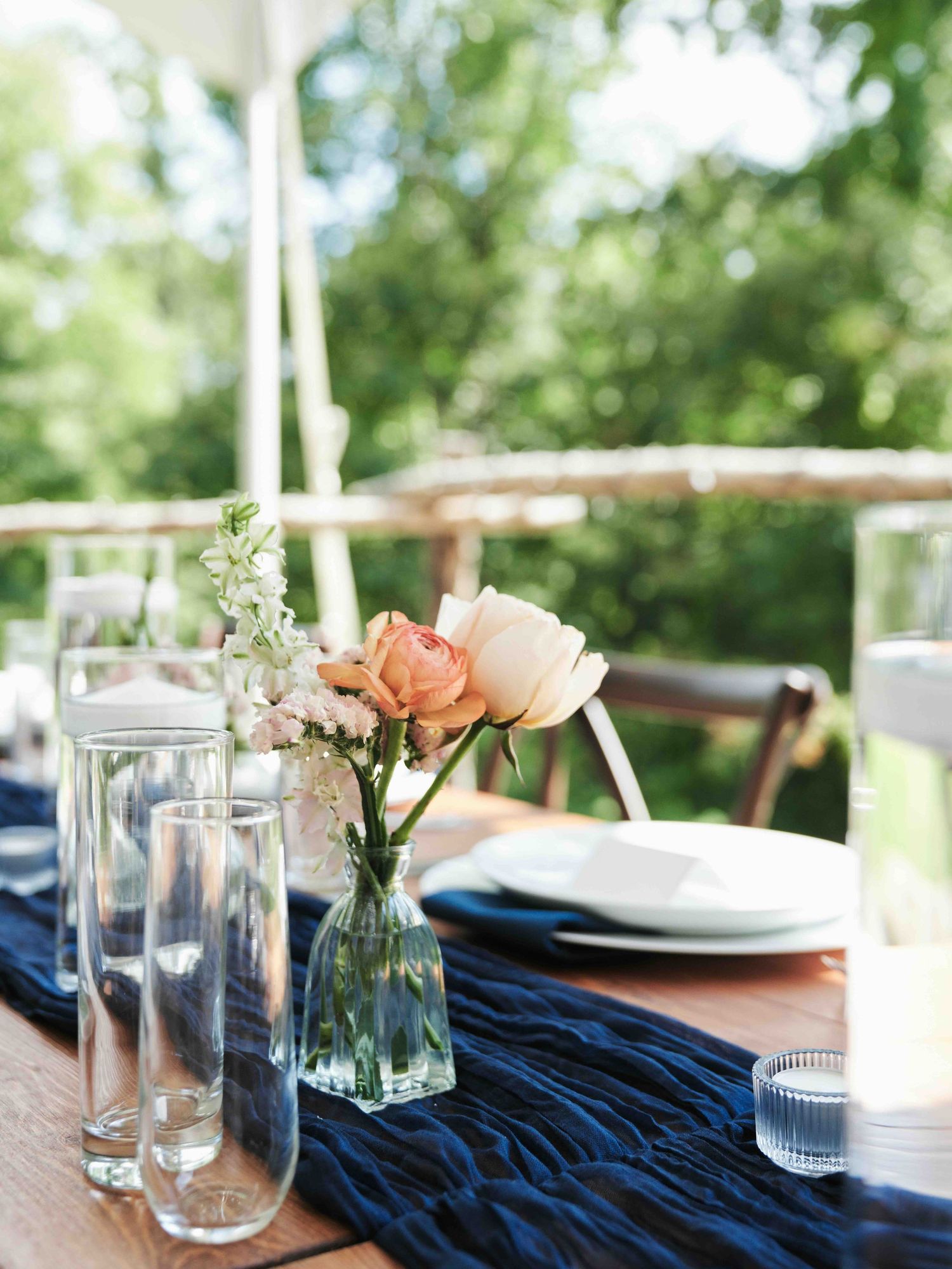 Navy blue table runner with peach roses in glass vases on a wooden table at an outdoor wedding reception.