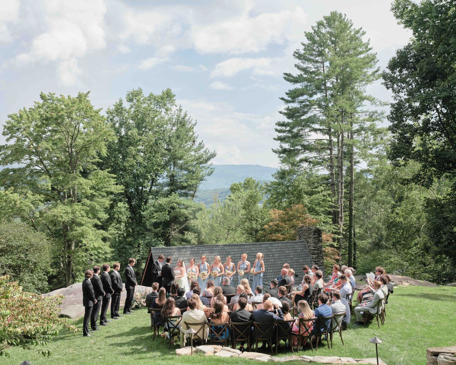 Outdoor wedding ceremony on a lush green hillside with scenic mountain views, surrounded by tall trees and seated guests.